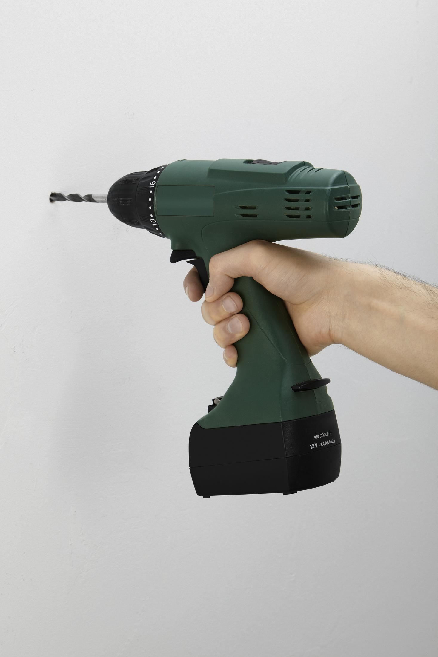 La mano de una persona sosteniendo un taladro eléctrico verde y negro. El taladro está contra una pared blanca.