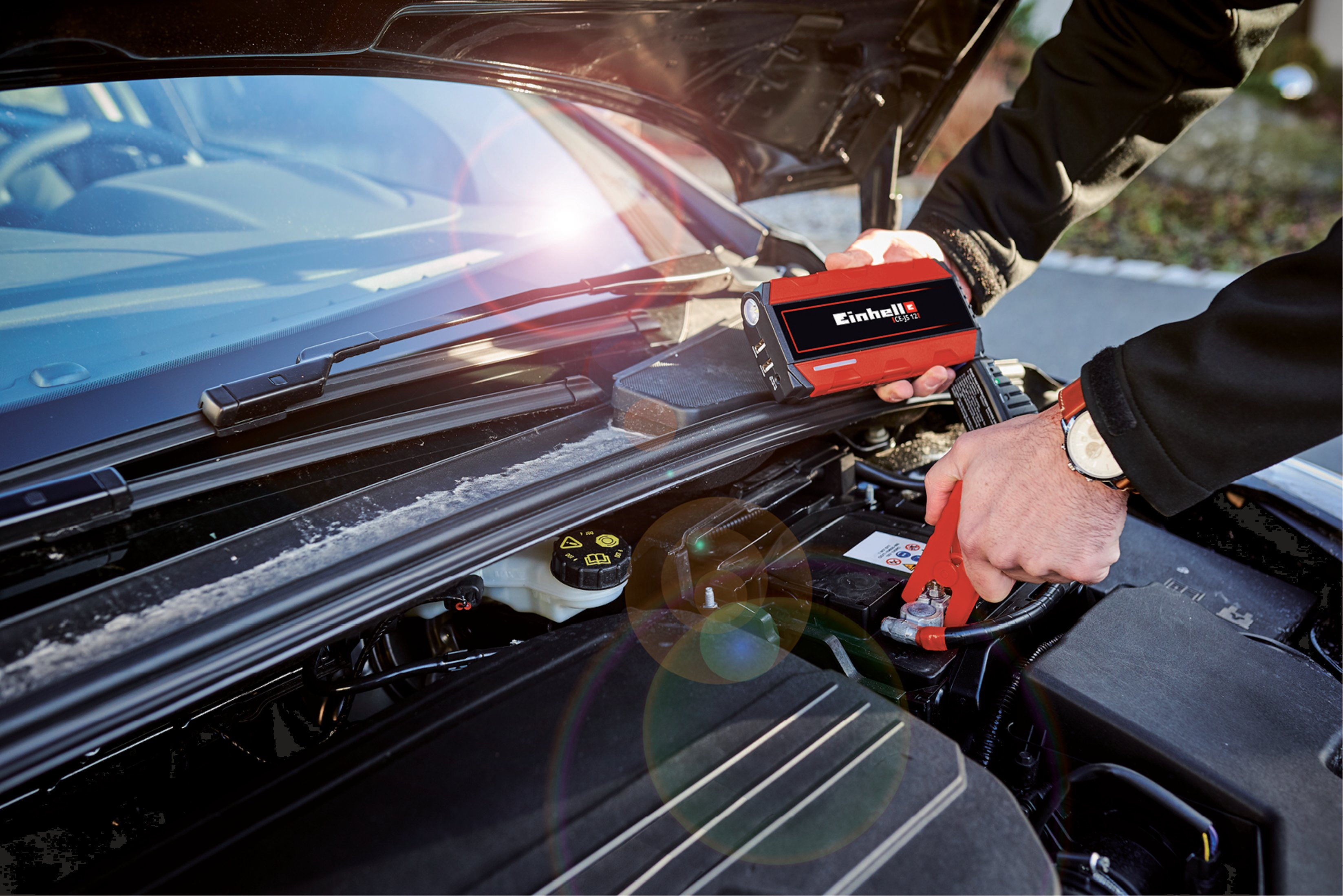 Ein Mann verbindet rote Überbrückungskabel mit einer Autobatterie im Motorraum.
