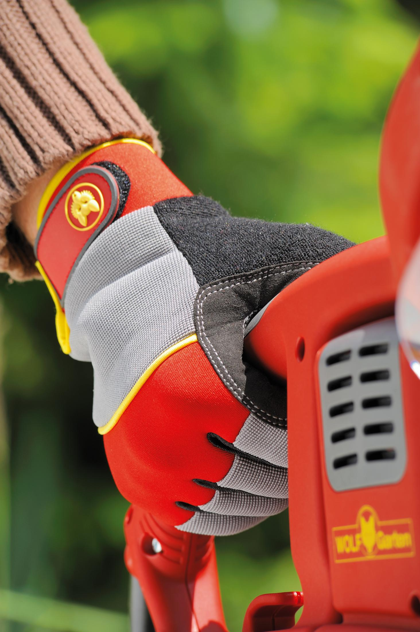 Eine behandschuhte Hand hält eine rote Wolf Garten Heckenschere. Der Handschuh ist rot, grau, gelb und schwarz.
