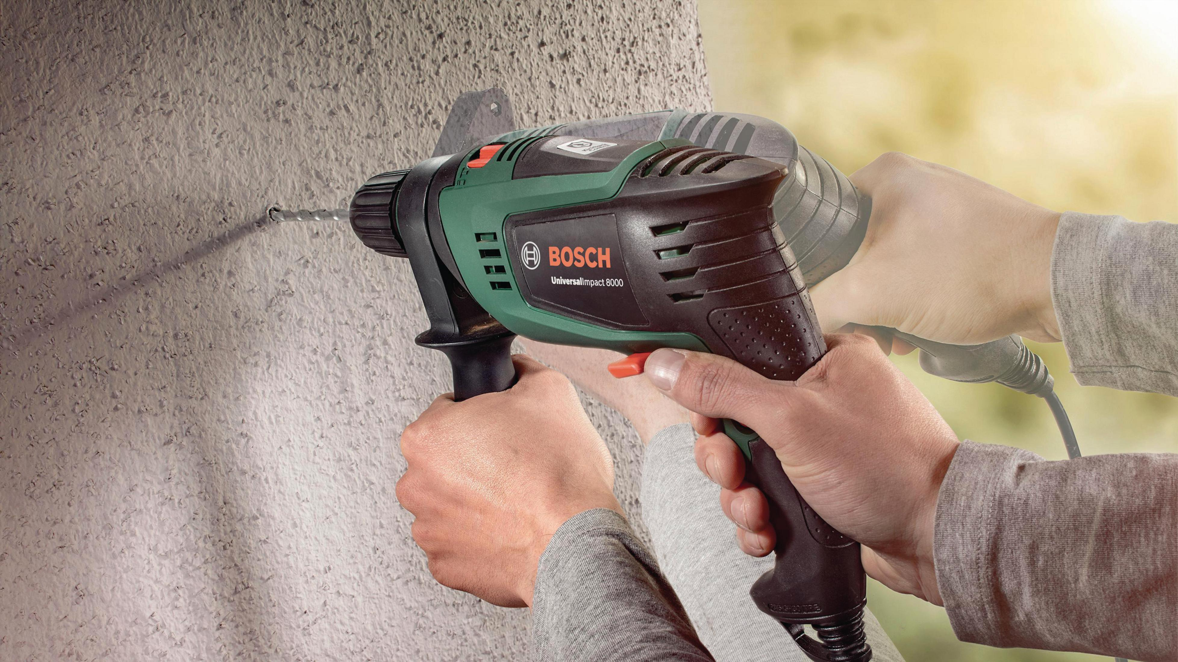Bir kişi, beton duvara delmek için bir Bosch darbeli matkap kullanıyor. Eller görünür.