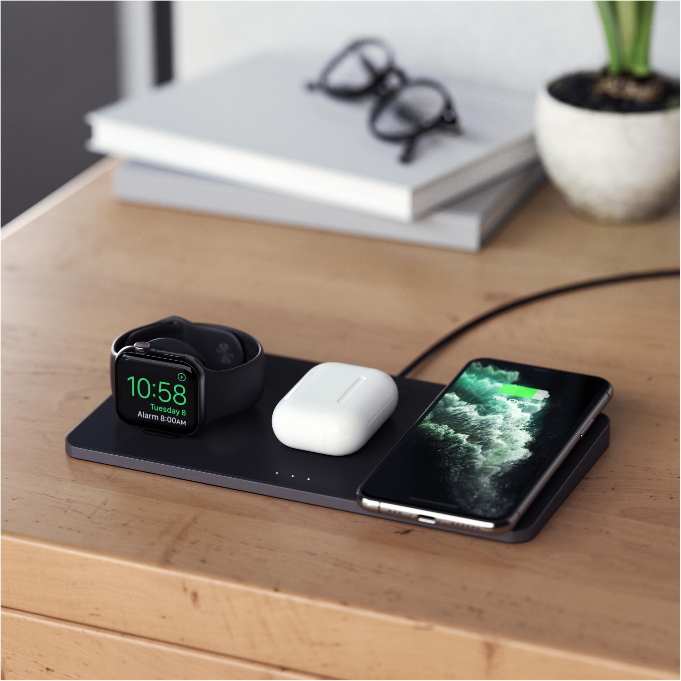 Una estación de carga con un reloj negro, auriculares blancos y un teléfono, todo sobre una mesa de madera.