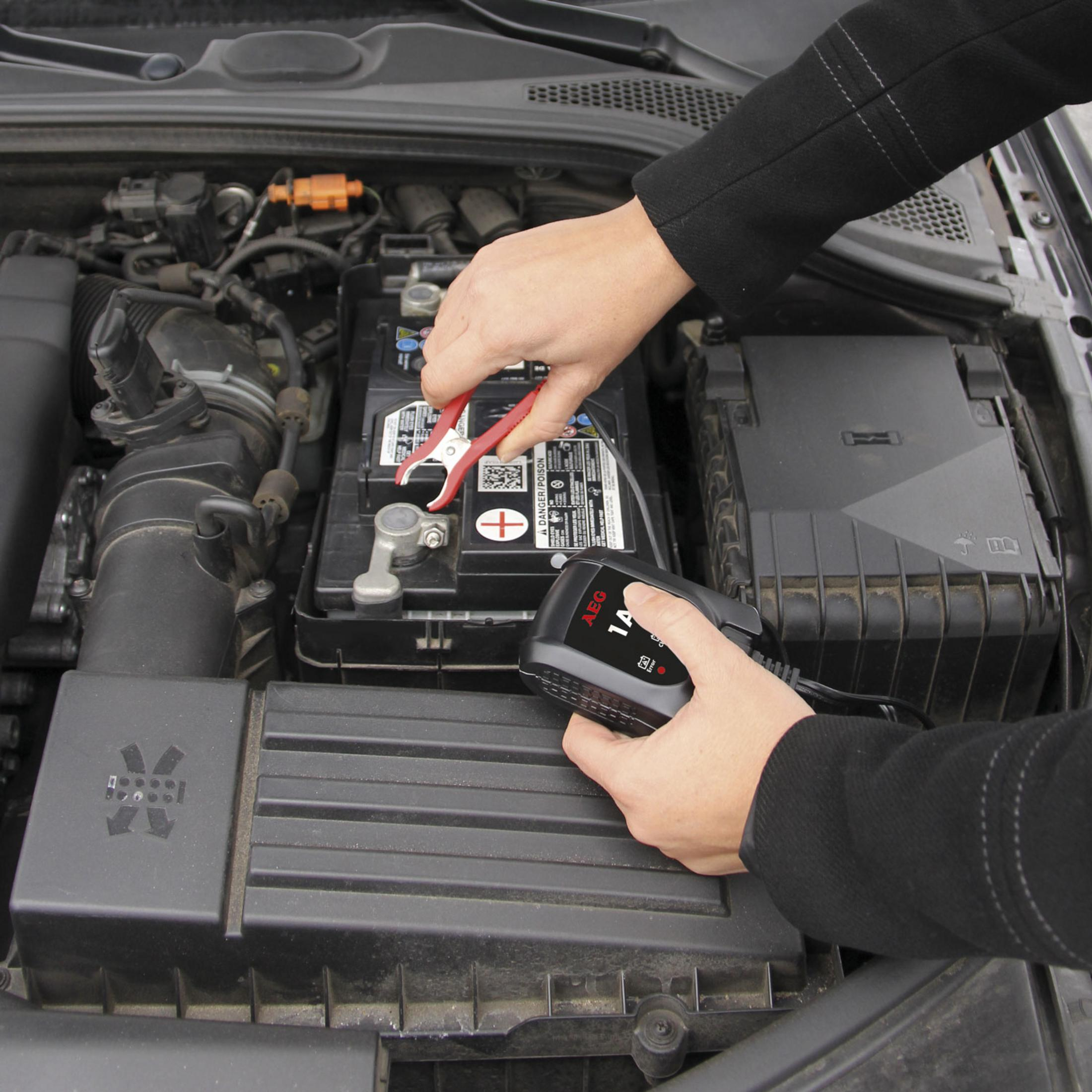 Hände, die eine rote Zange an einer Autobatterie verwenden, Ladegerät daneben.