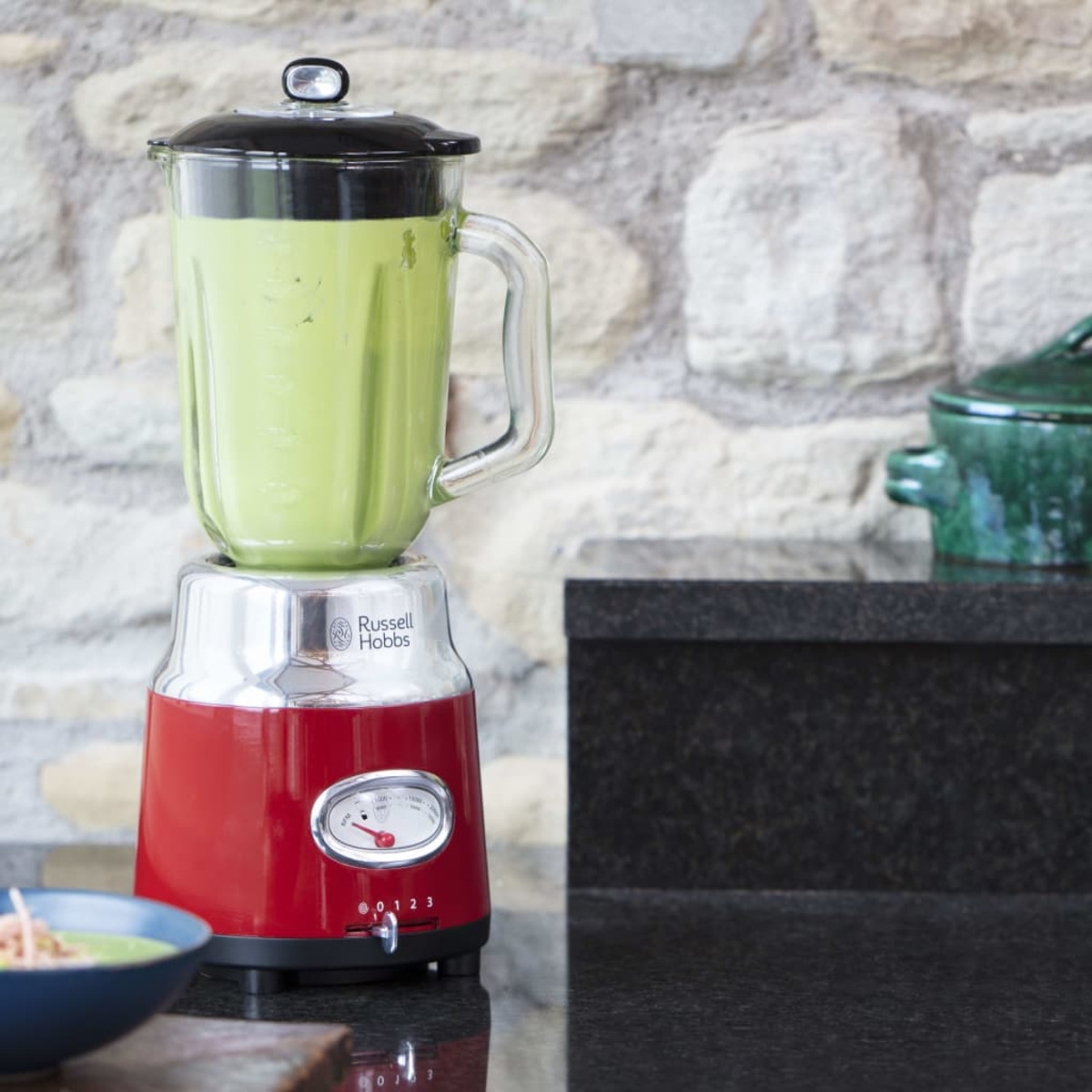 Kırmızı ve gümüş renkli, yeşil sıvılı bir blender. Blender'ı bir tezgahta gösterir.