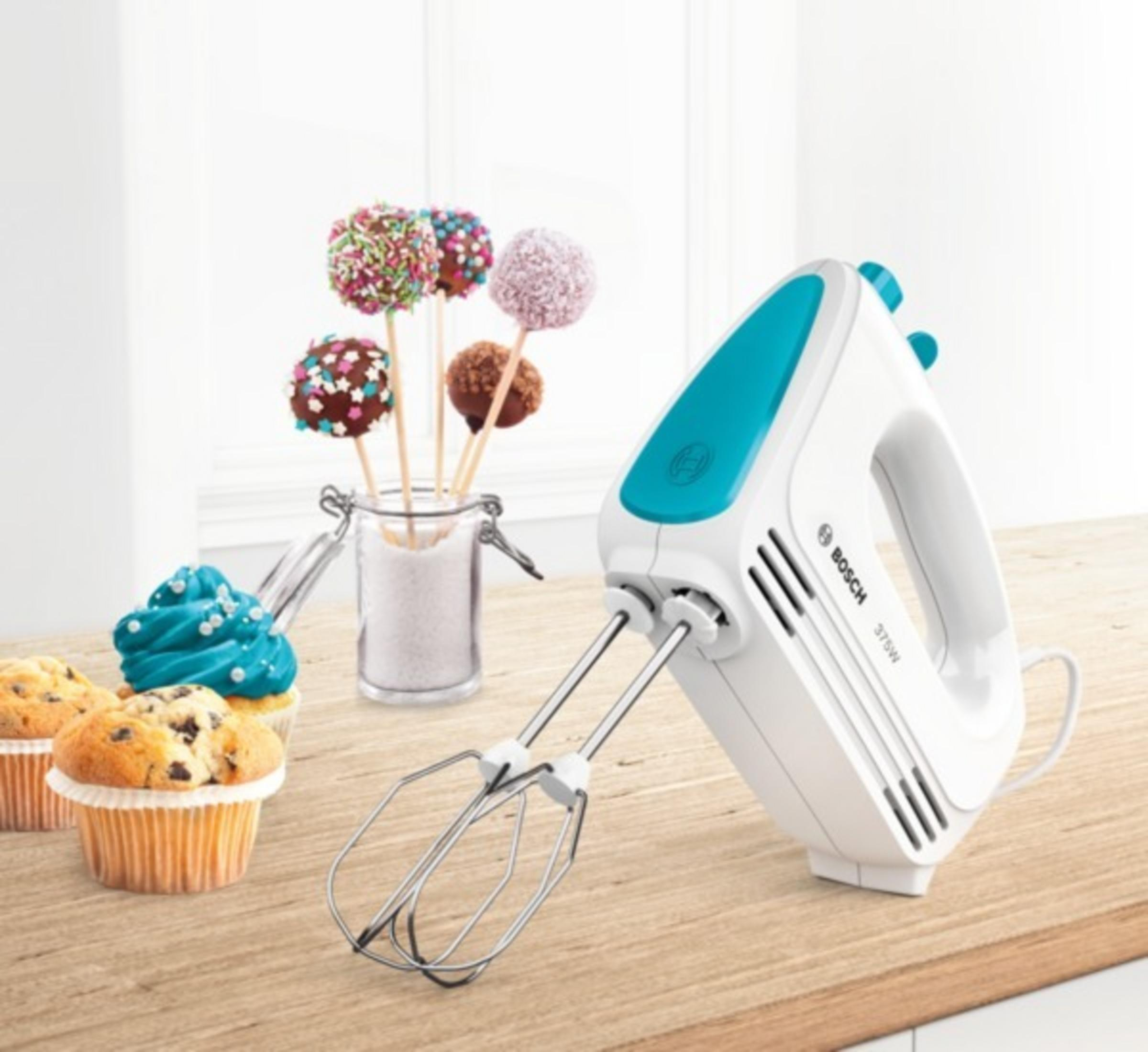 A white and teal hand mixer with cupcakes and cake pops on a wooden surface.
