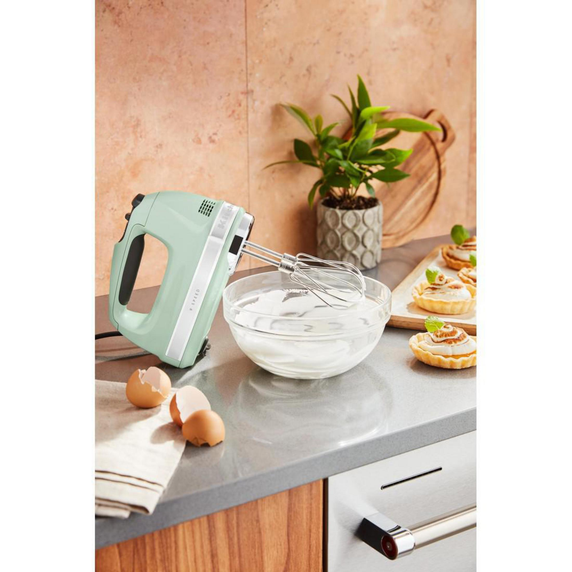 Minzgrüner Handmixer in Glasschüssel. Eier und Torten daneben. Küchenszene.