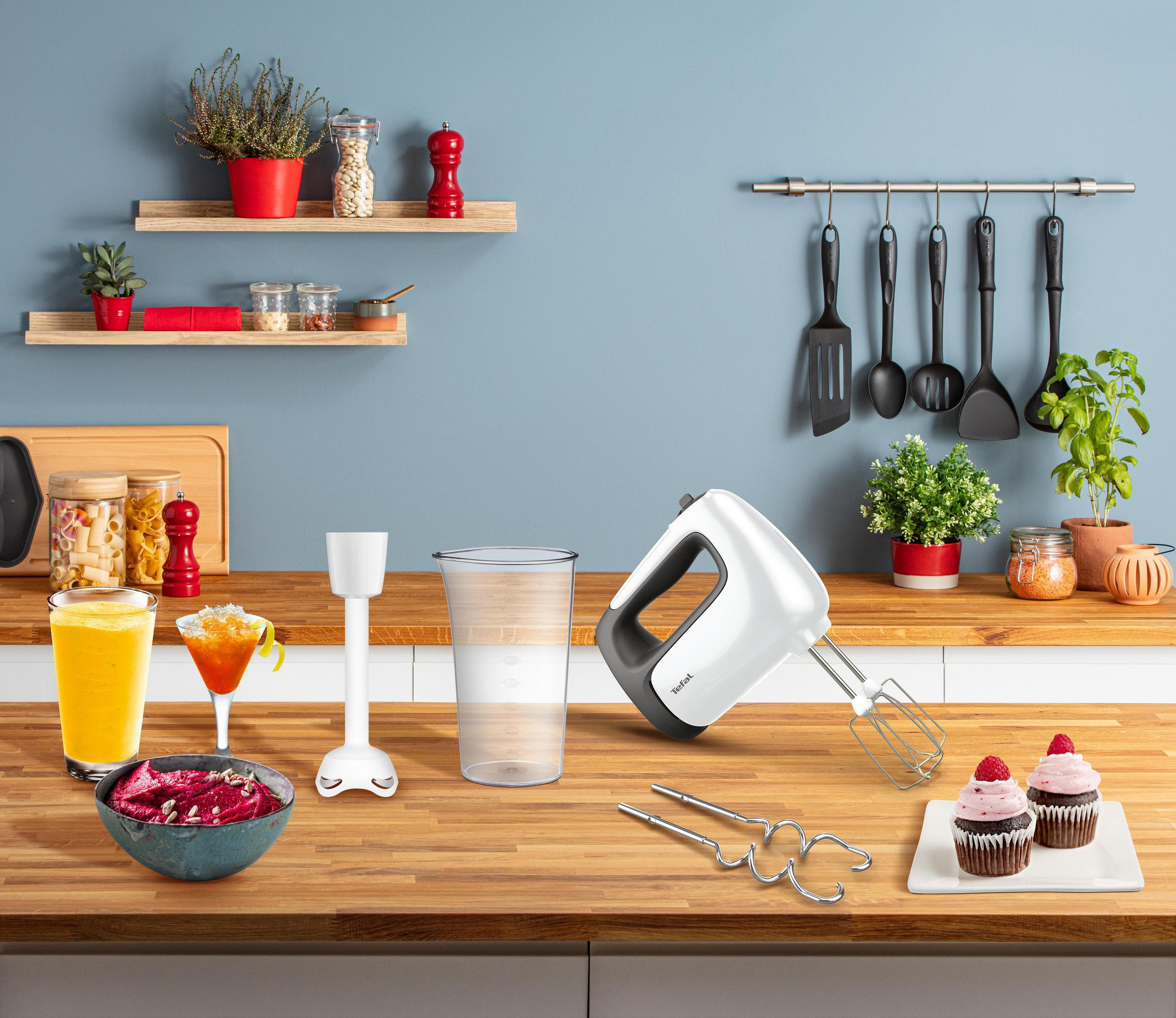 Scena kuchenna: drewniany blat z blenderem, mikserem, szklankami, babeczkami i półkami z roślinami.