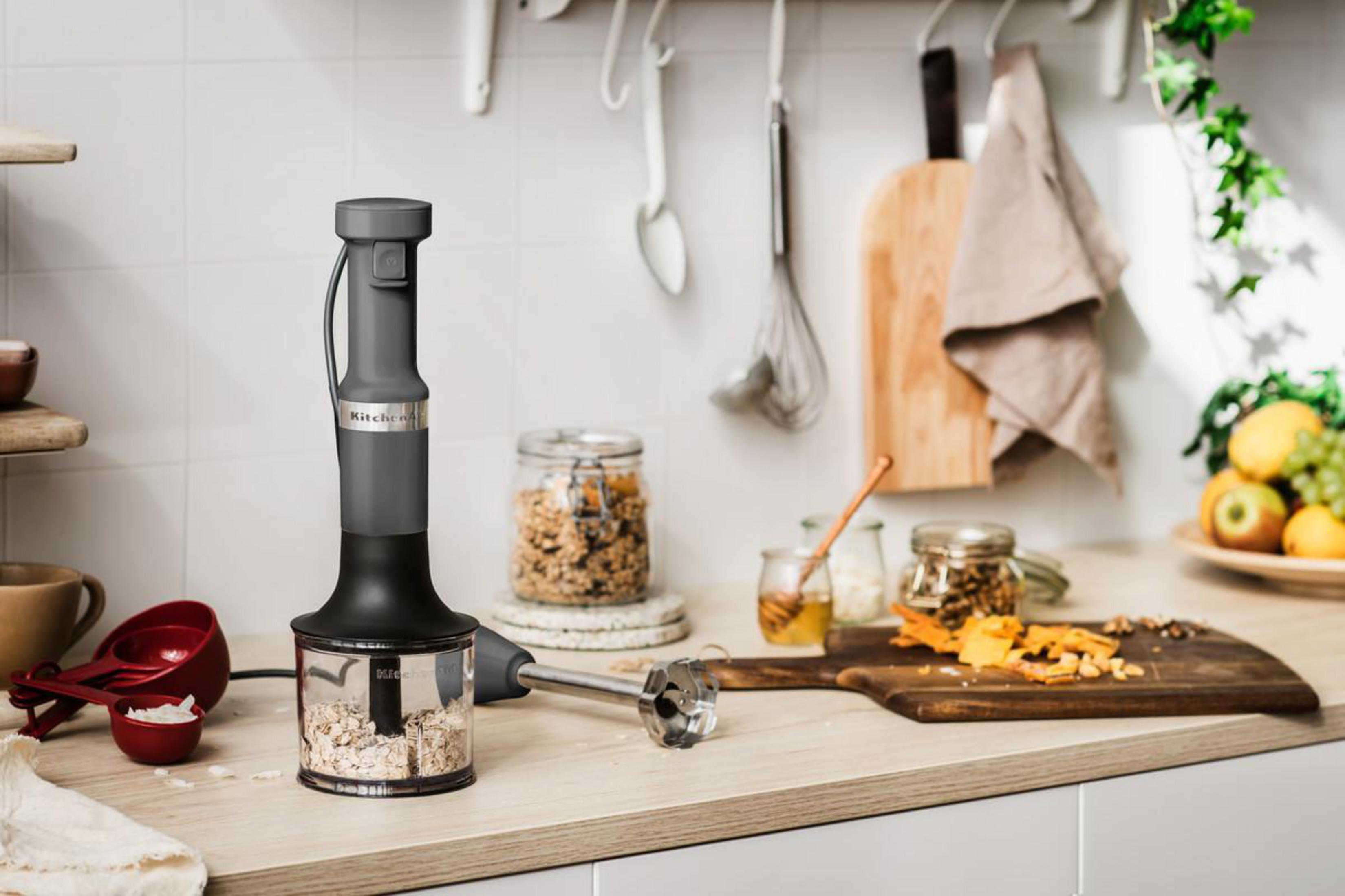 Escena de cocina con una batidora de inmersión Kitchenaid en uso. Otros elementos incluyen frascos, tablas de cortar y fruta.