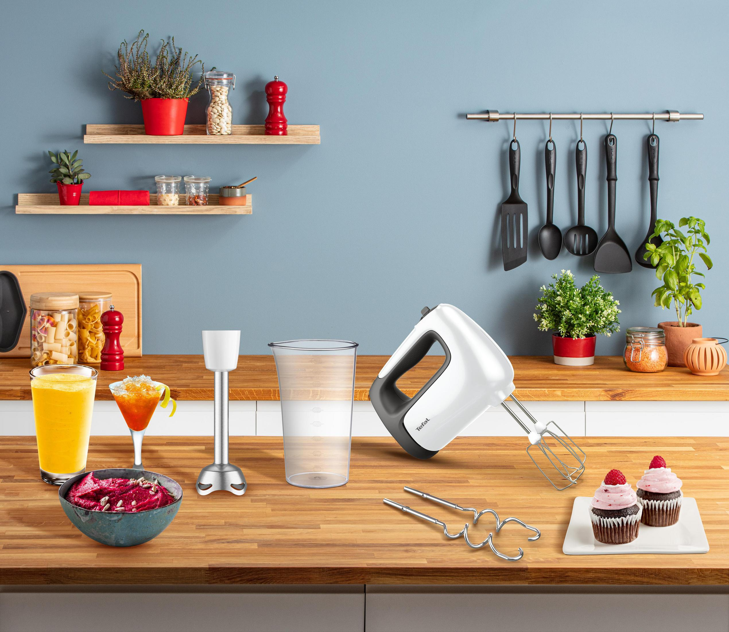 A kitchen counter with food, a blender, and cooking utensils in front of a blue wall.