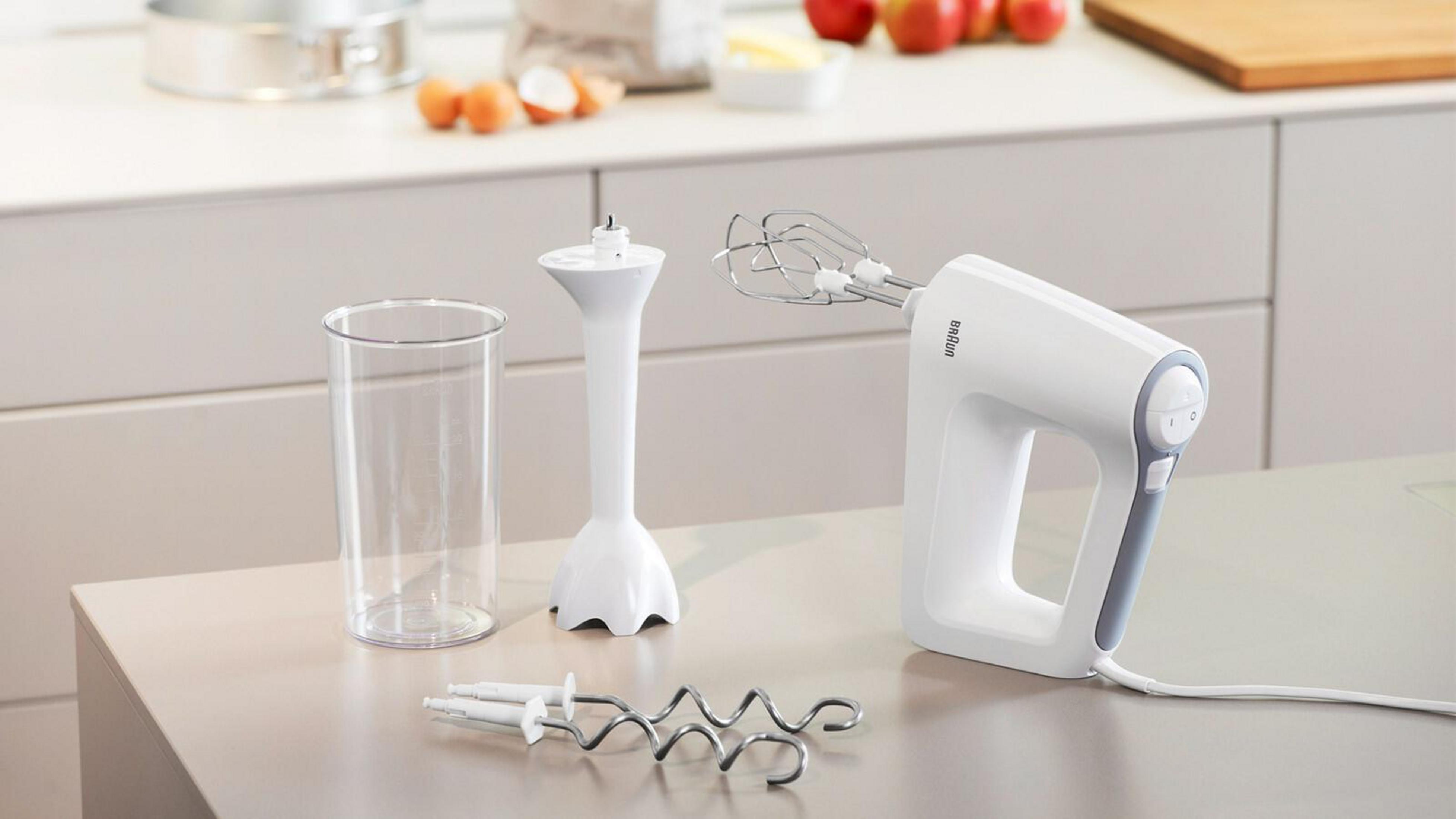 Weißer Handmixer mit Zubehör und einem Glas. Backzutaten im Hintergrund.