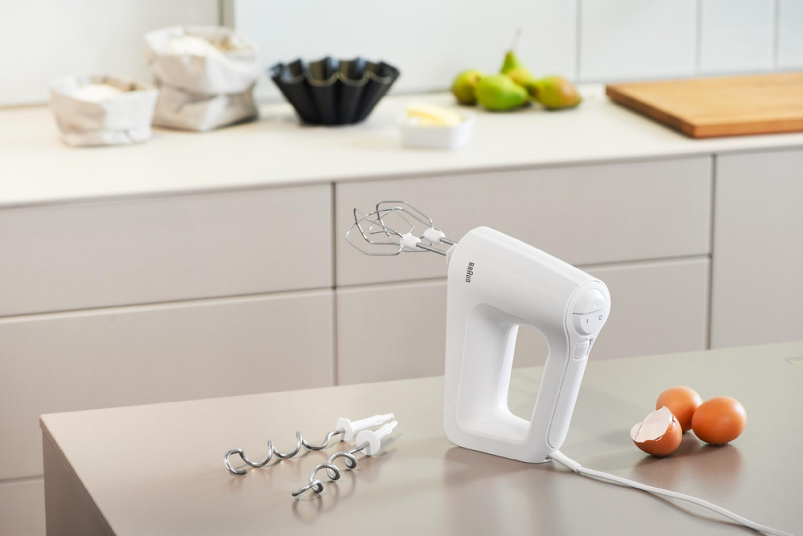Weißer Handmixer mit Aufsätzen auf Metalloberfläche. Eier, eine Schüssel und Zutaten im Hintergrund.