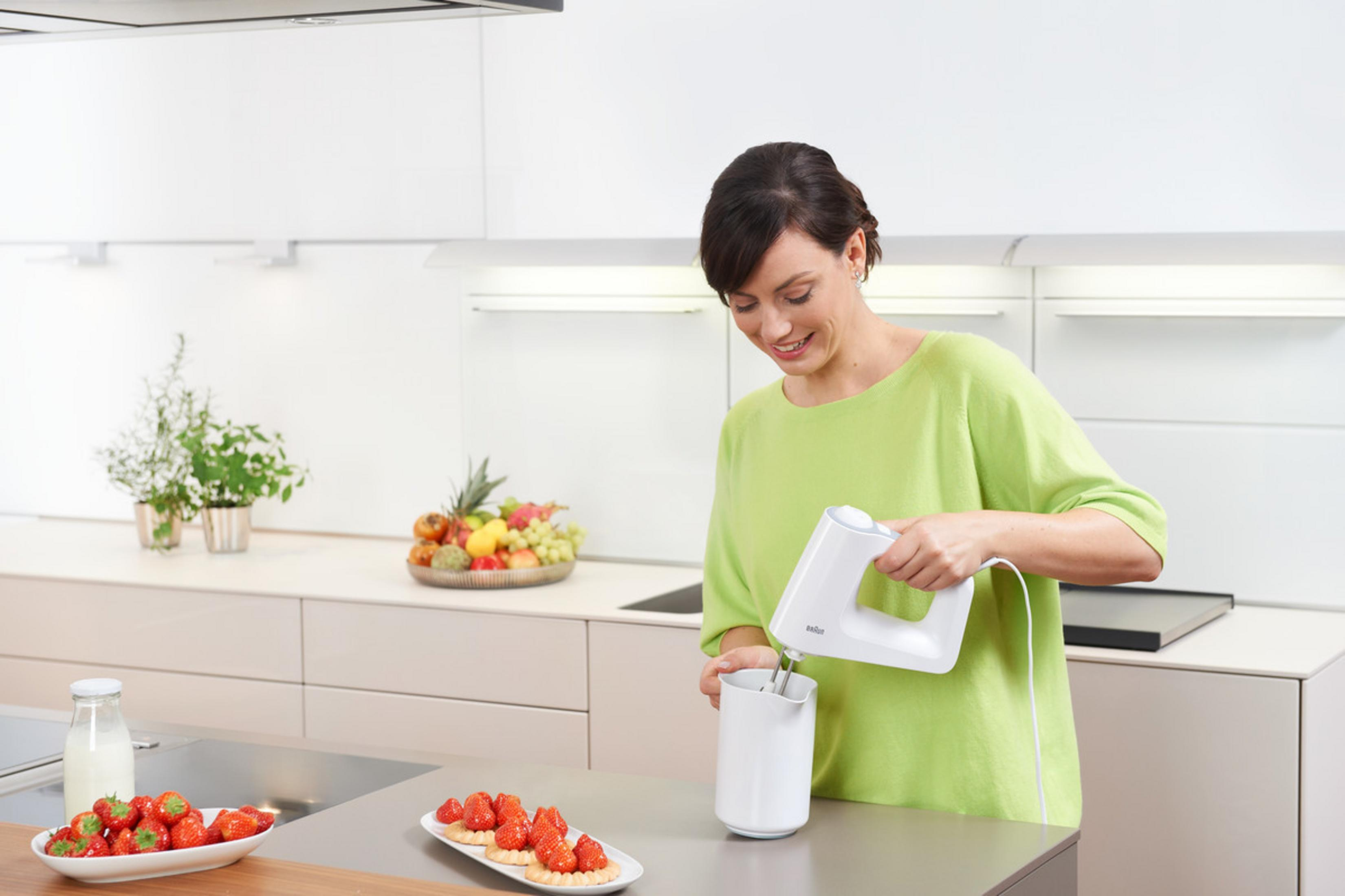 Frau in grünem Oberteil, die einen weißen Handmixer benutzt. Erdbeeren und Milch auf dem Tisch, Küche im Hintergrund.