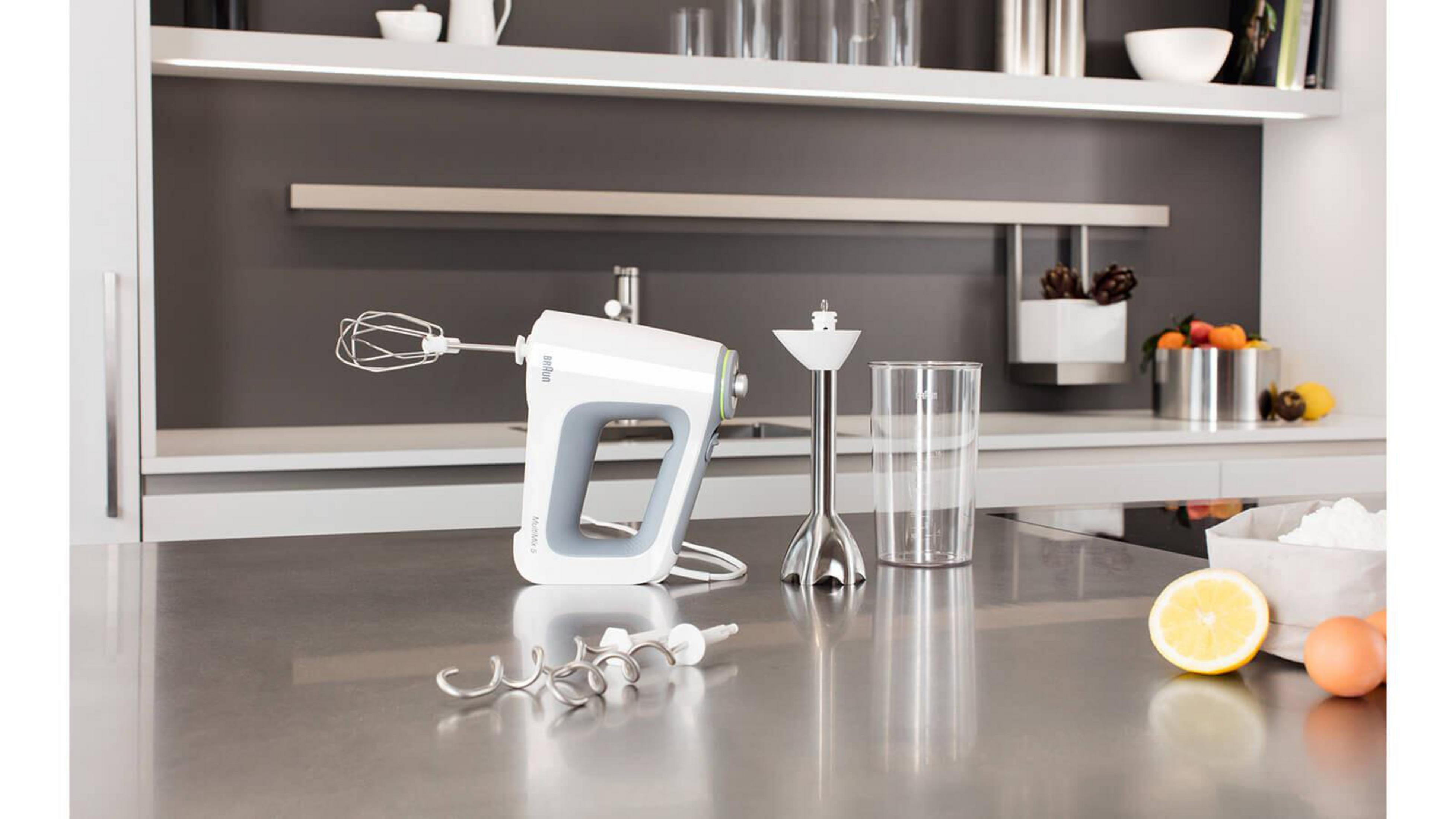 Ein weißer Handmixer mit Aufsätzen, ein Mixer und ein Glas. Alle stehen auf einer Theke.