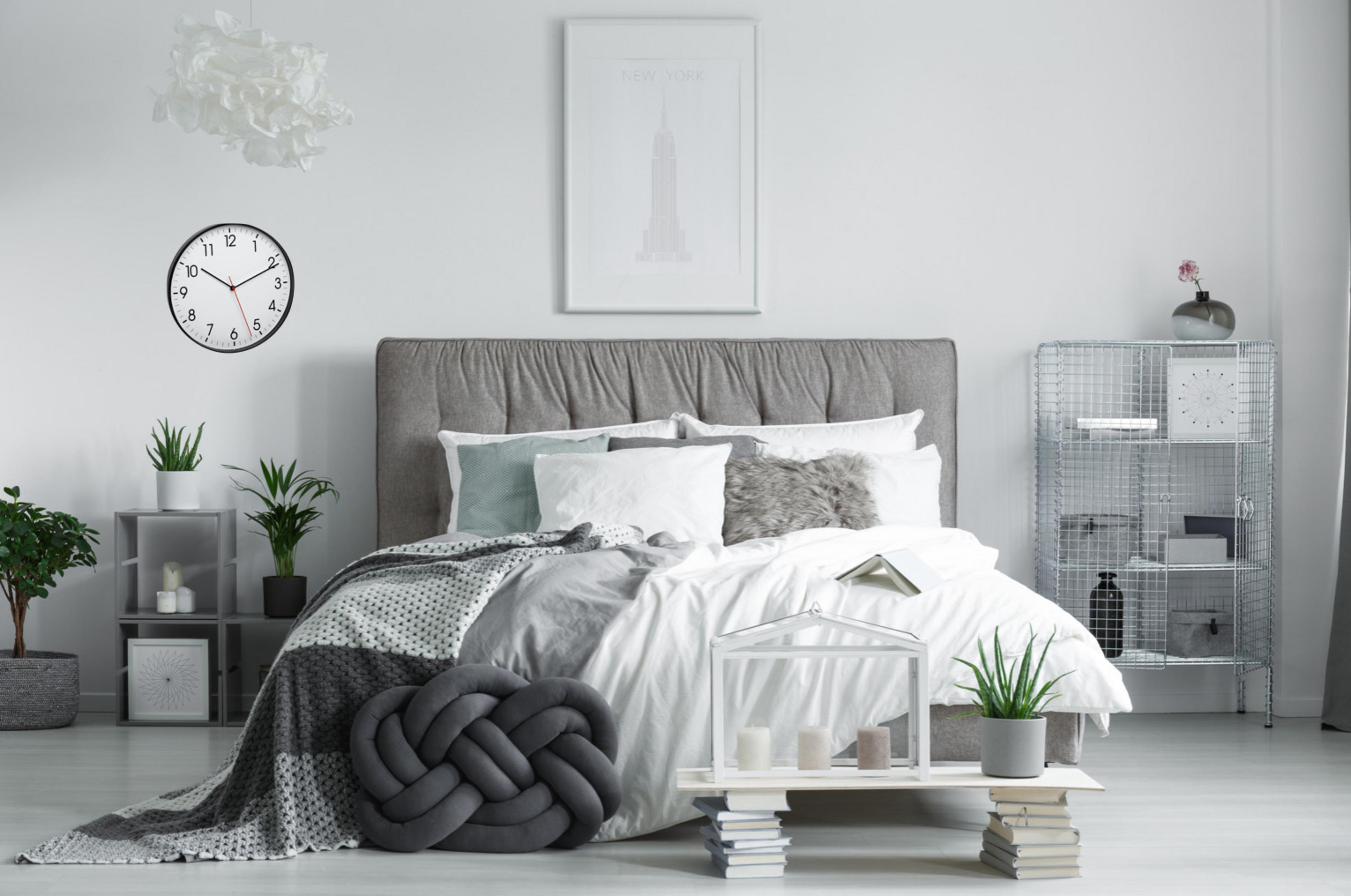 Schlafzimmer-Szene. Bett mit grauem Kopfteil, weißer Bettwäsche, grauer Decke, Deko, Uhr, Pflanzen und New York Bild.