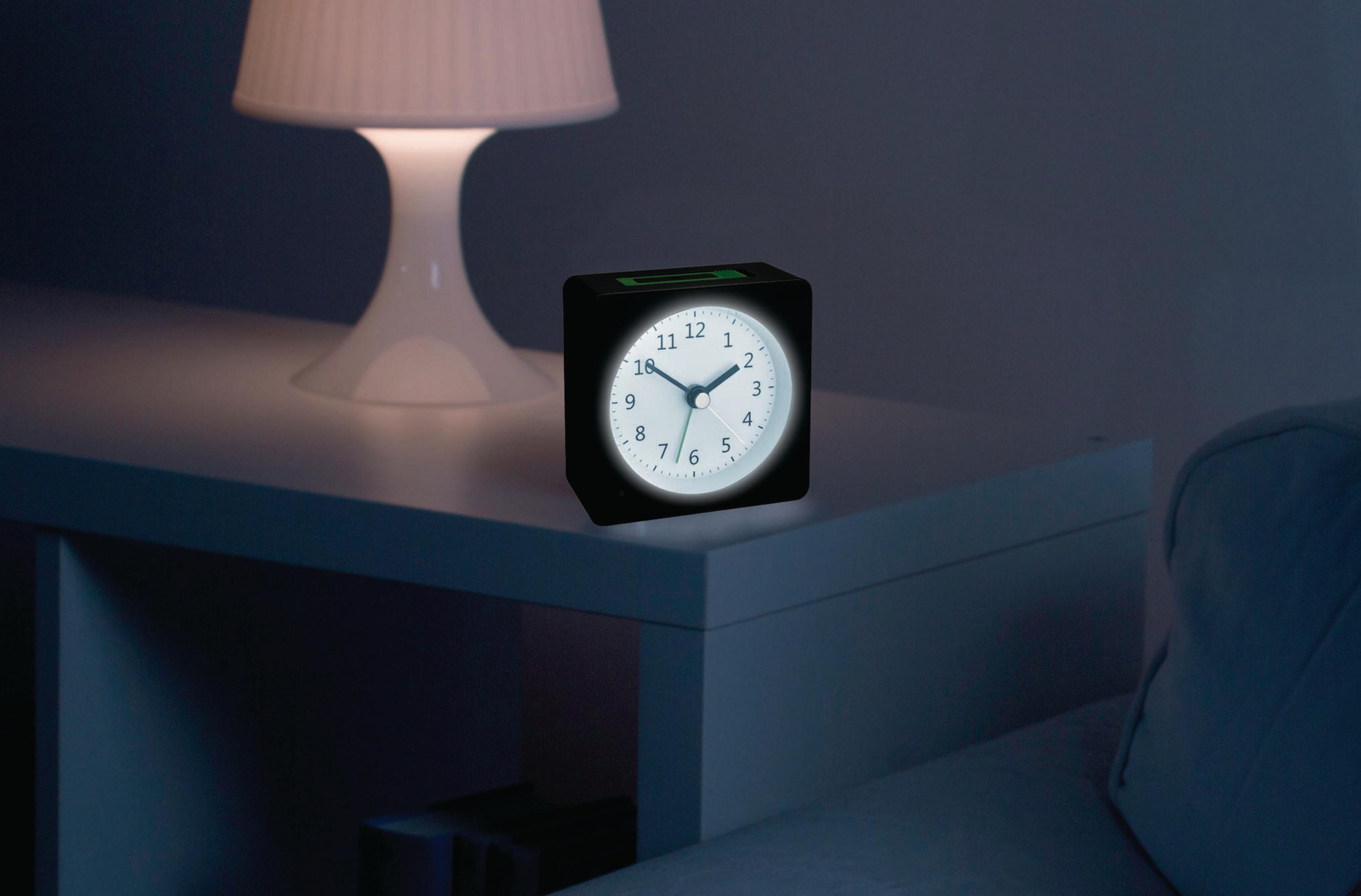 Schwarzer Wecker auf einem weißen Tisch neben einer Lampe und einem Sofa.