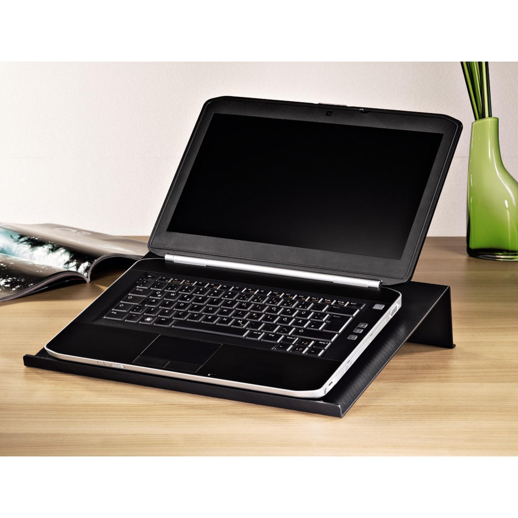 Laptop nero su un supporto nero. Tastiera visibile. Tavolo in legno, vaso verde, libro aperto, parete bianca.