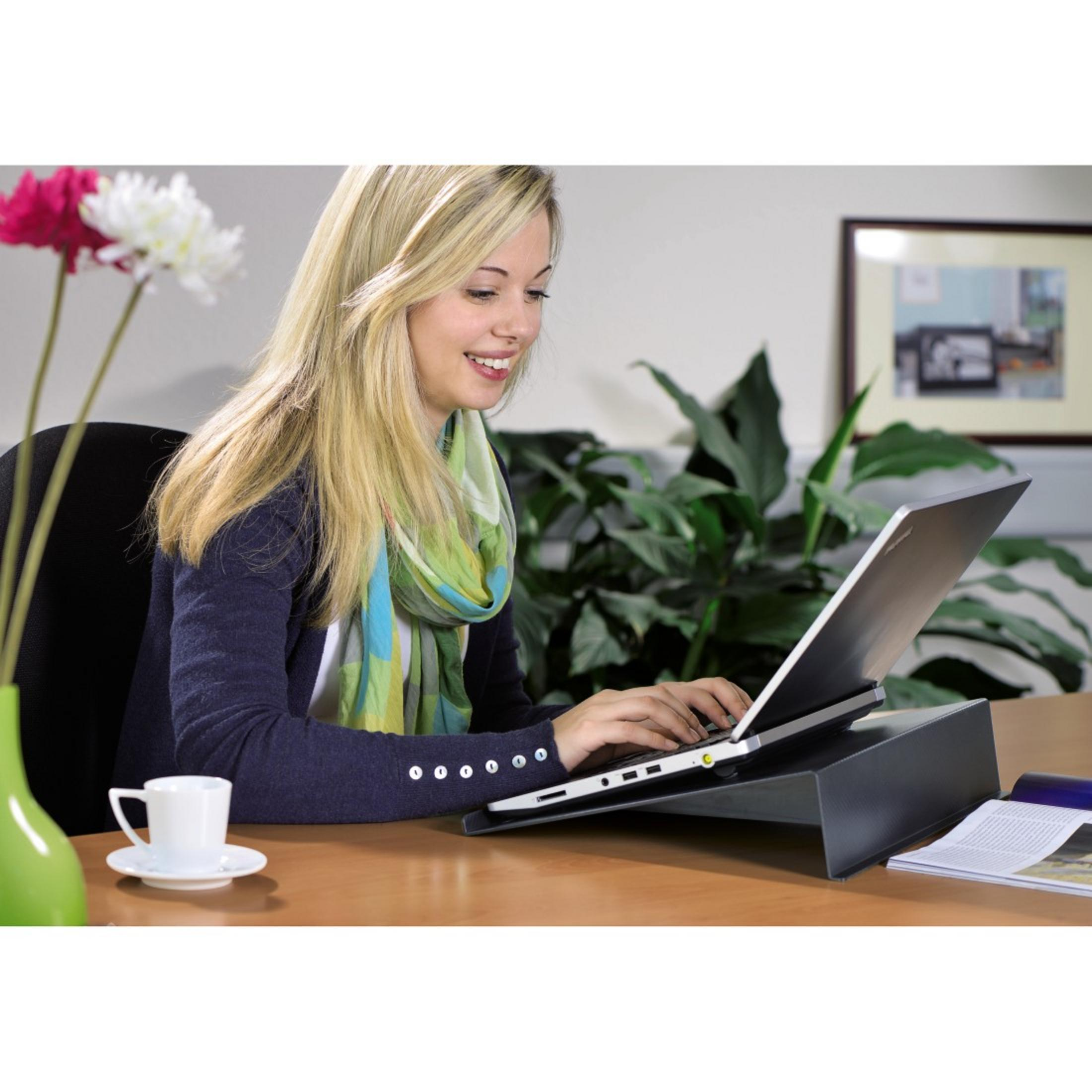Donna bionda che lavora su un laptop. È seduta a una scrivania con una tazza di caffè e fiori. Laptop su un supporto.