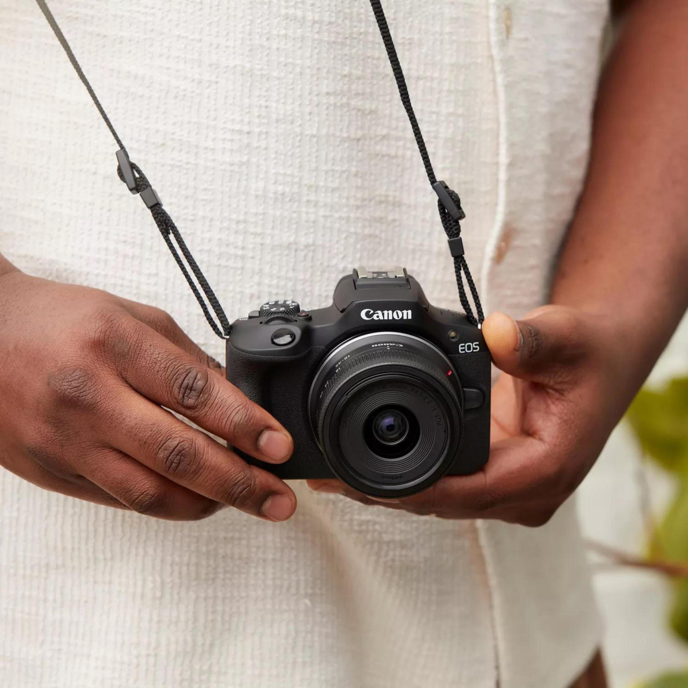 A person holding a black Canon EOS camera with a strap, against a light shirt.