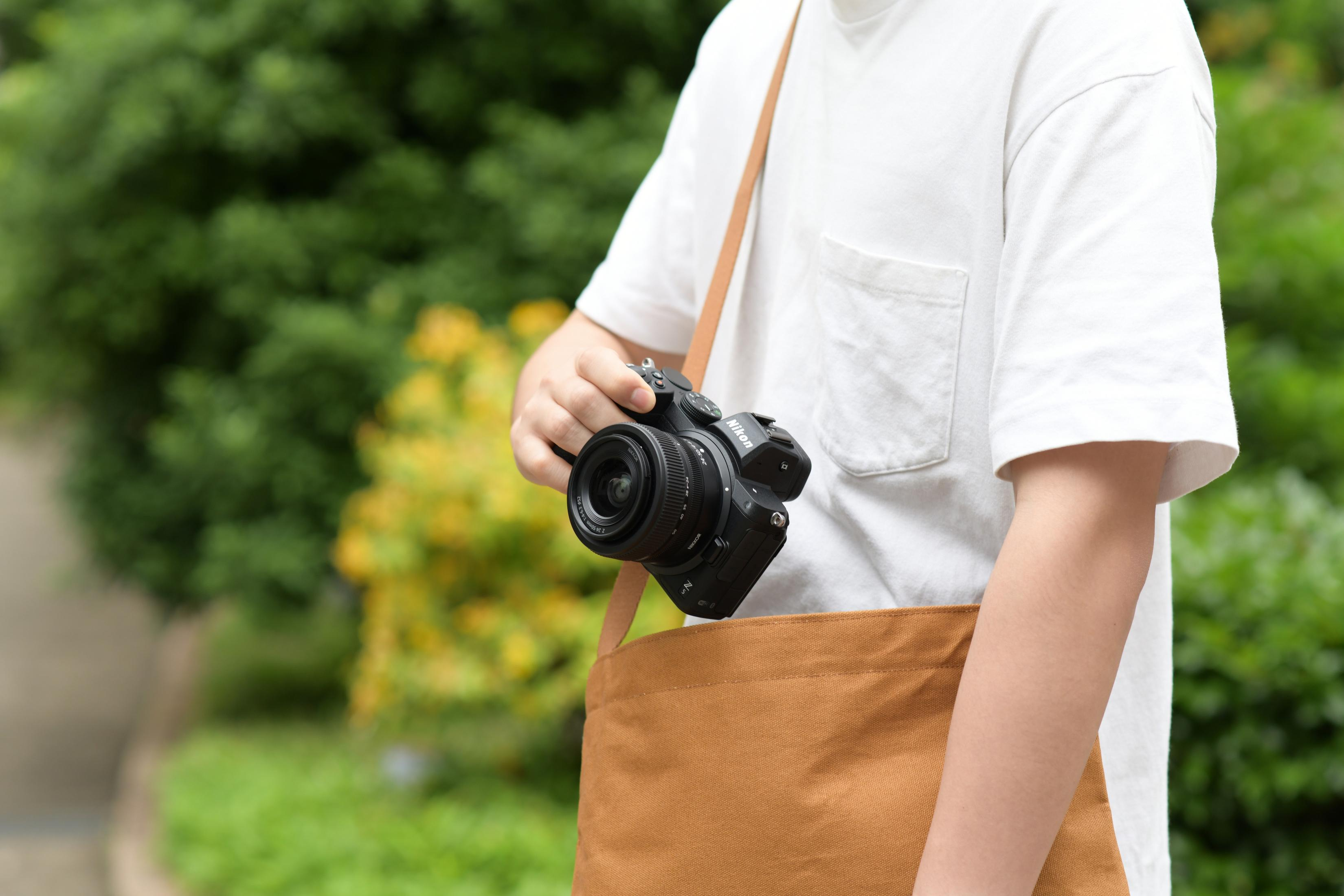 Person hält eine schwarze Kamera mit brauner Tasche über der Schulter. Grüner Hintergrund.