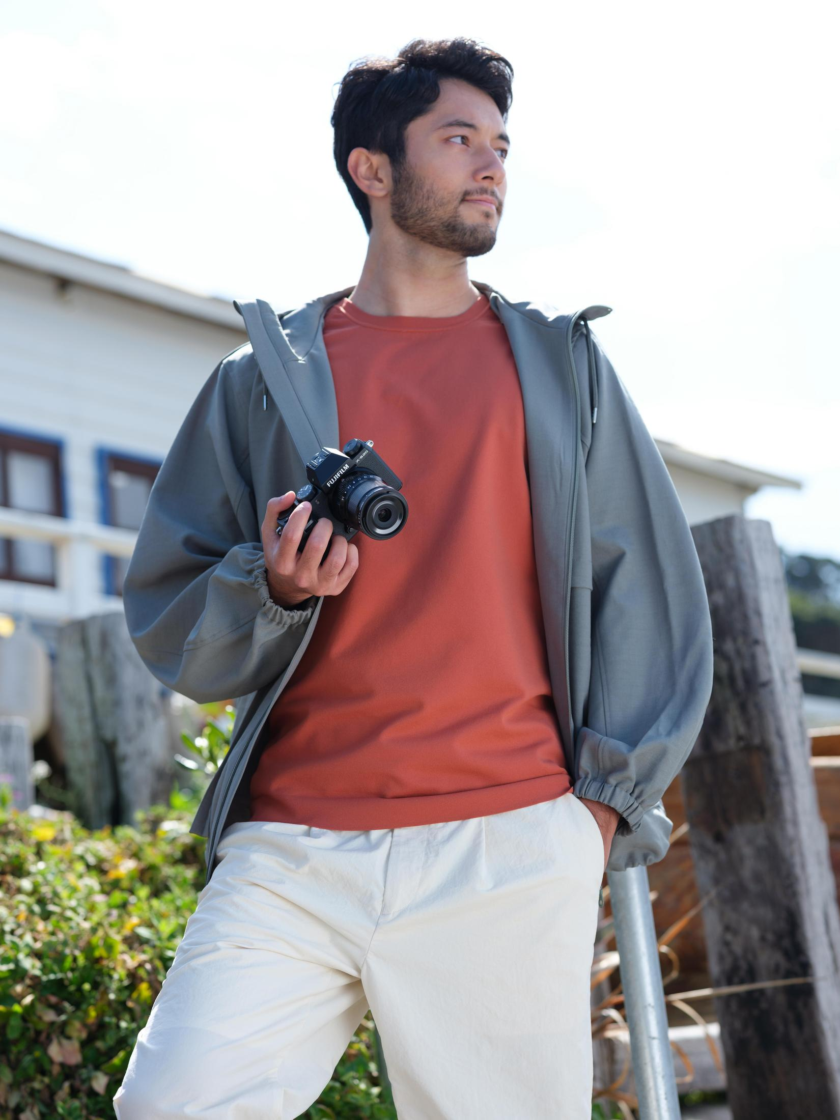 Un uomo con giacca grigia e camicia arancione che tiene una fotocamera. Lo sfondo è una giornata di sole.
