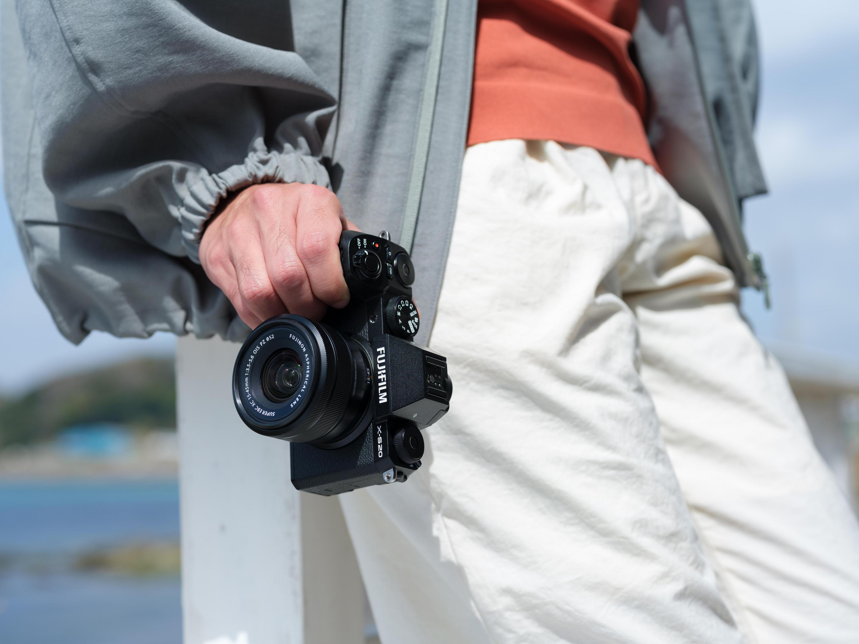 Persona che tiene una fotocamera Fujifilm nera, con cielo blu e acqua sullo sfondo.