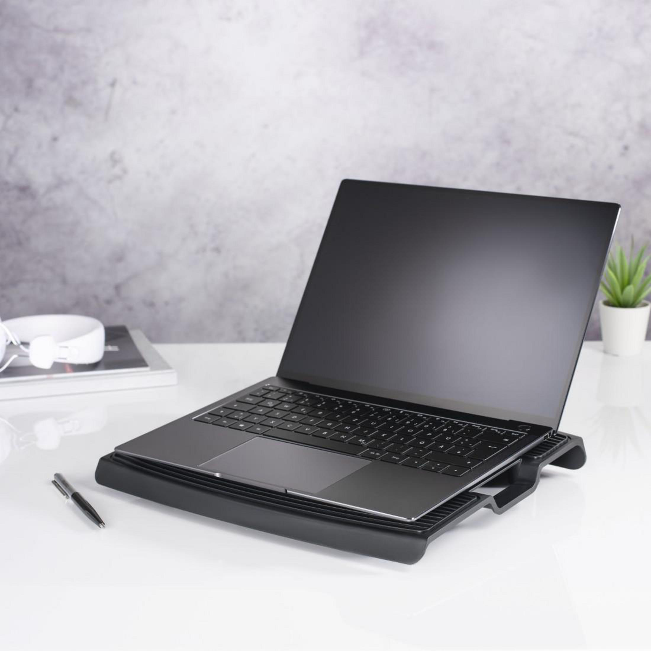 Laptop auf schwarzem Ständer, Stift, Kopfhörer und Buch auf weißem Tisch, graue Wand.