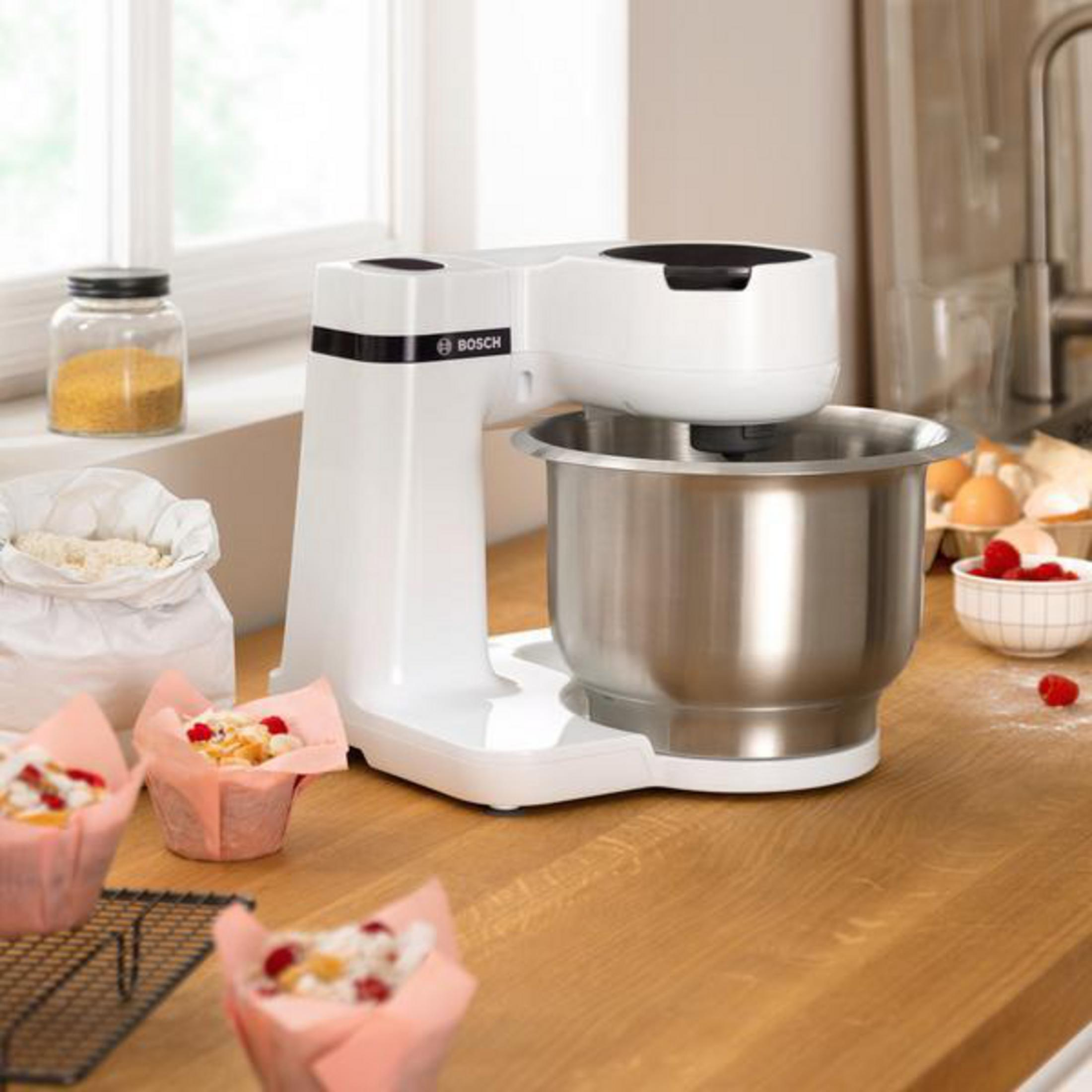 Eine weiße Küchenmaschine mit silberner Schüssel, Cupcakes und Zutaten auf einem Holztisch.