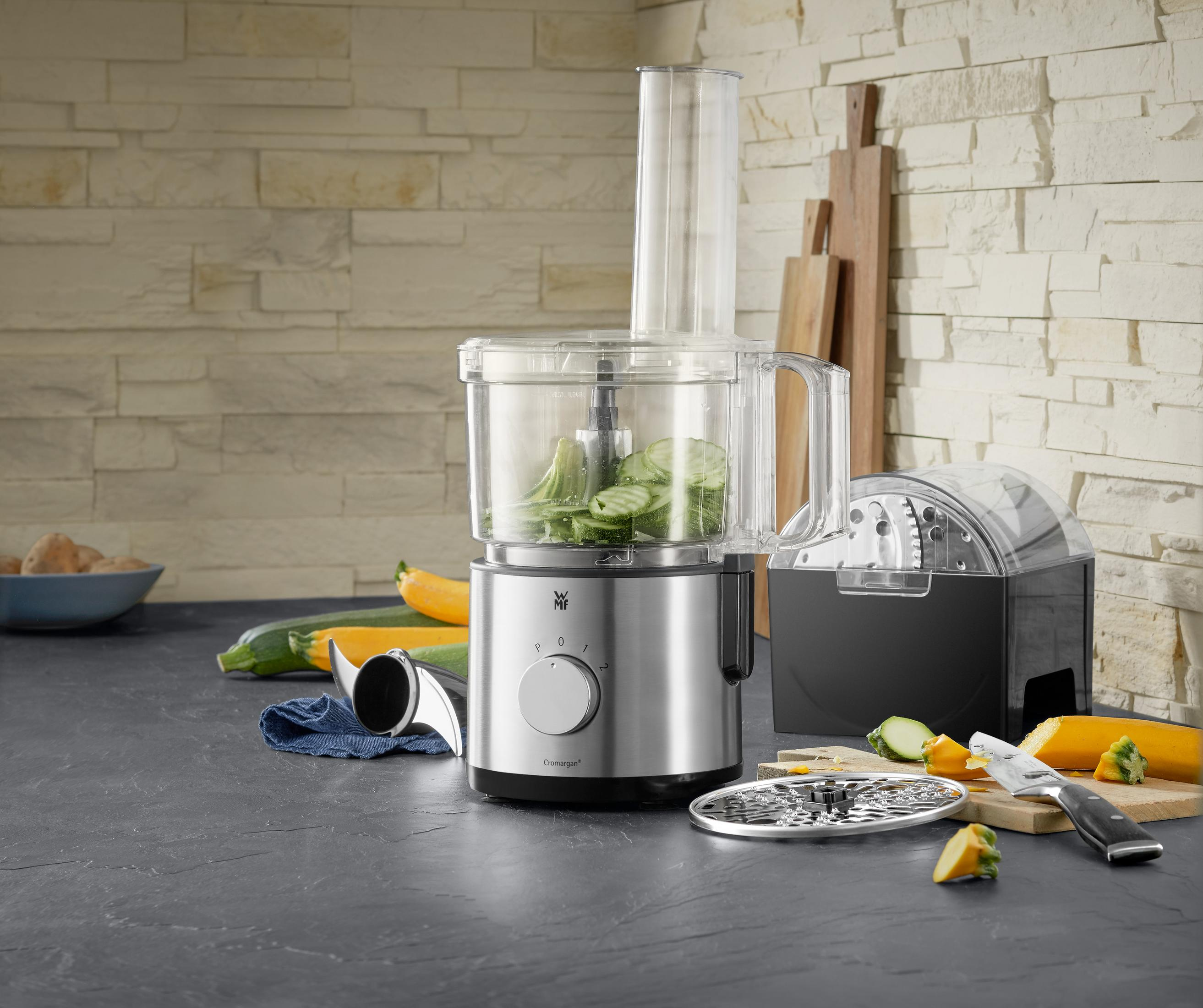 Un robot da cucina argentato e nero. Ha un contenitore trasparente con verdure all'interno, con accessori.