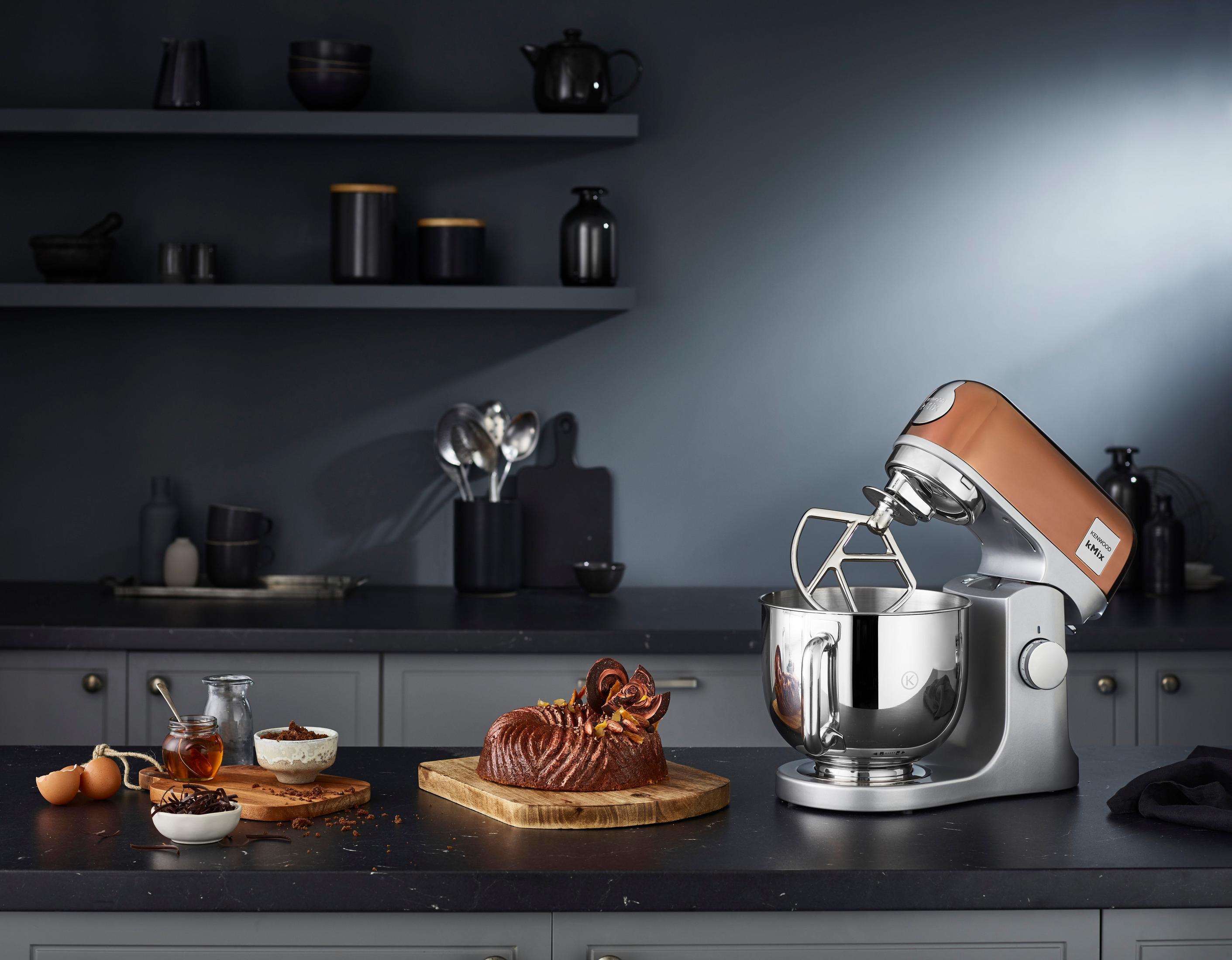 Cucina con un robot da cucina in rame e ingredienti. Una torta su un tagliere. Tema scuro.