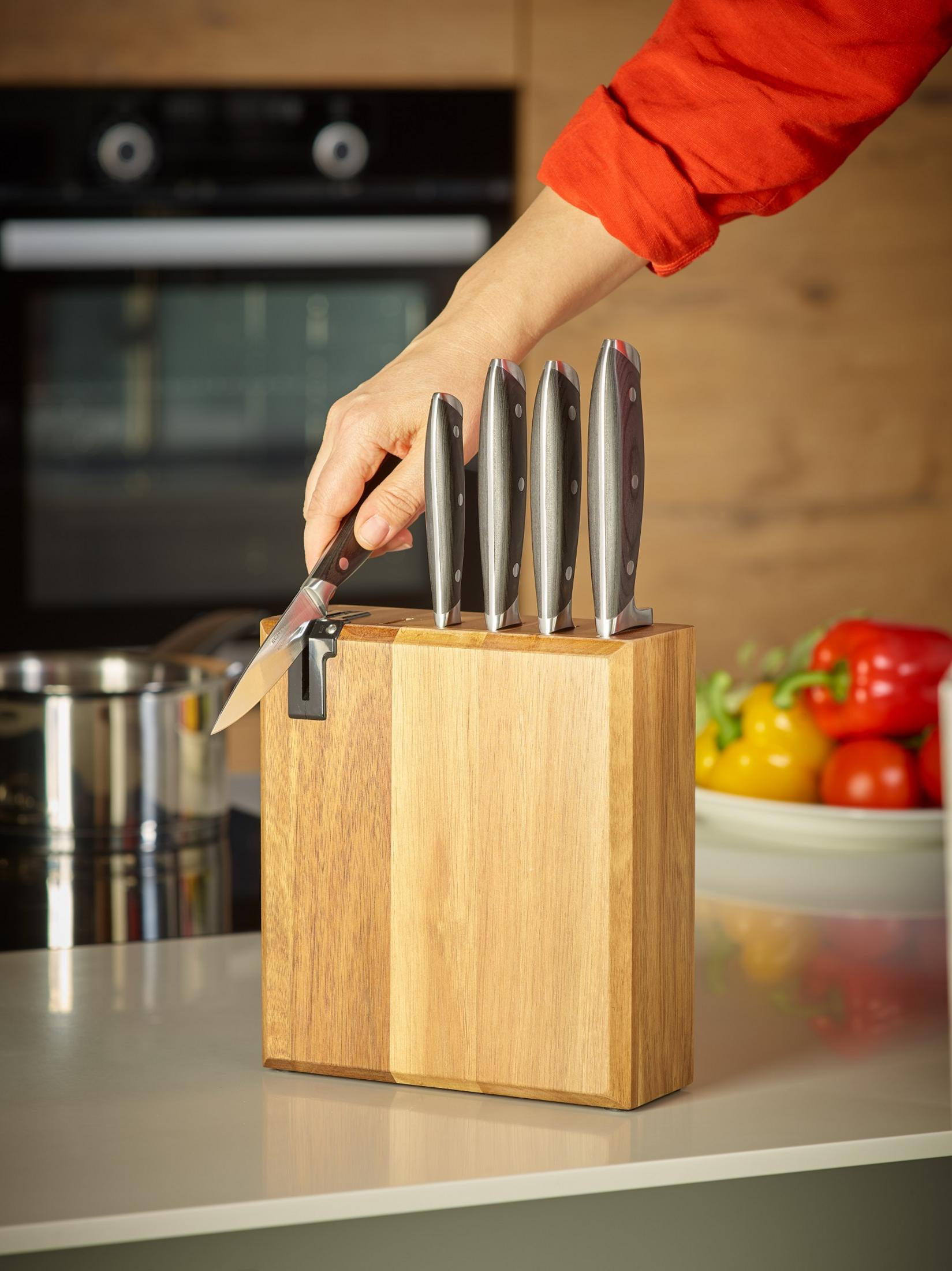 Eine Hand, die ein Messer in einen hölzernen Messerblock steckt, mit anderen Messern und Gemüse.