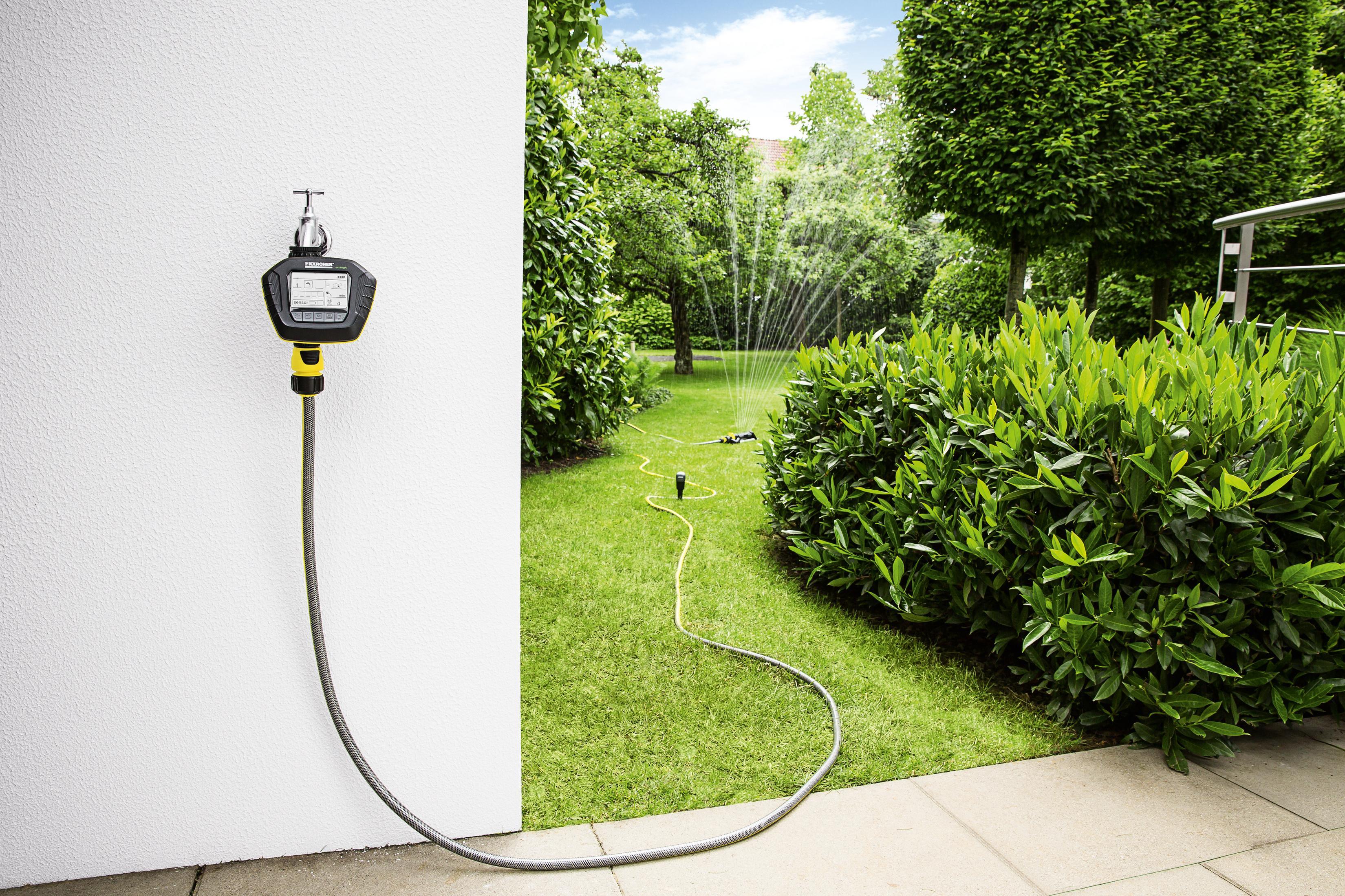 Ein Garten mit Sprinkleranlage und einem Wasser-Timer, der an einer Wand befestigt ist. Grüne Pflanzen und Gras.