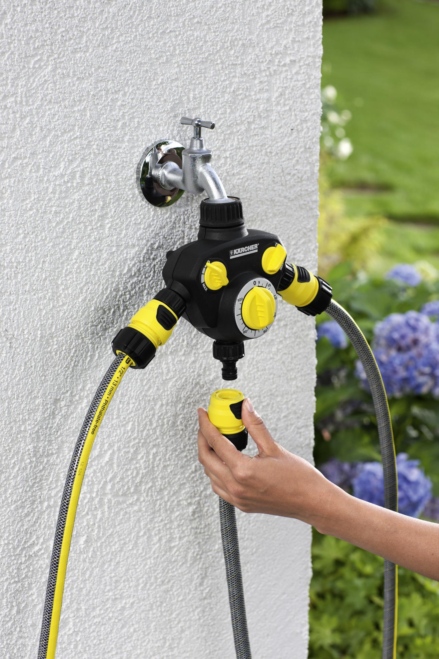 Ein Schlauch ist an einen schwarzen Wasserhahn mit Zeitschaltuhr angeschlossen, neben einer weißen Wand. Hand hält.