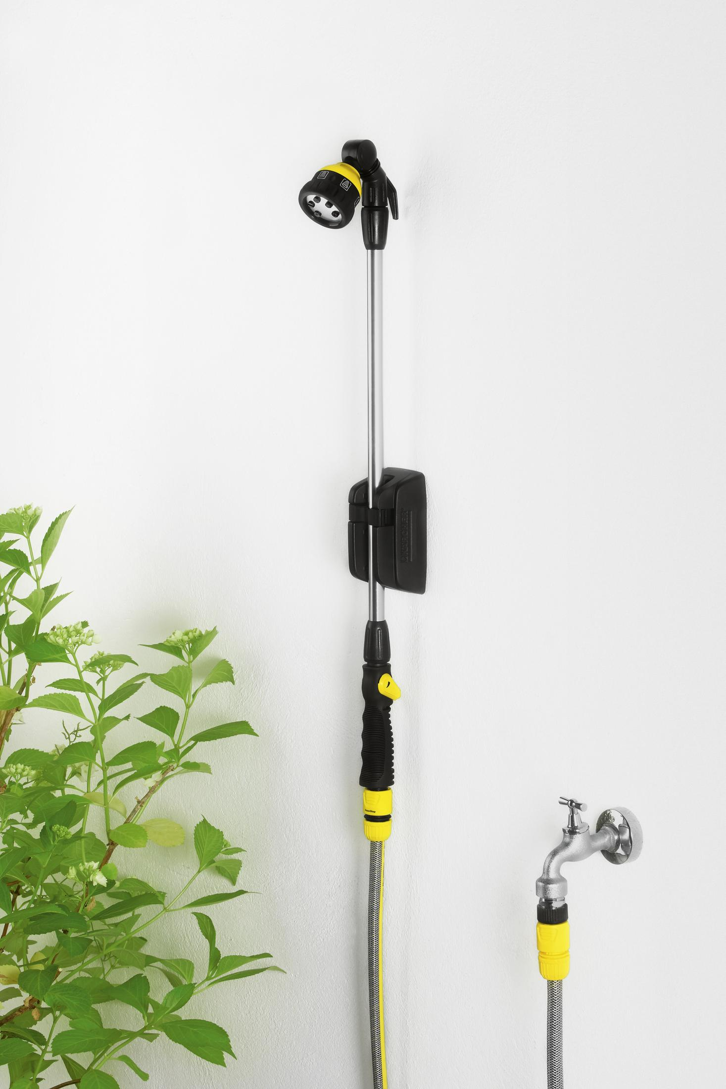 Eine Gartendusche an einer weißen Wand. Sie hat eine schwarze Halterung, gelbe Akzente und steht in der Nähe einer Pflanze.