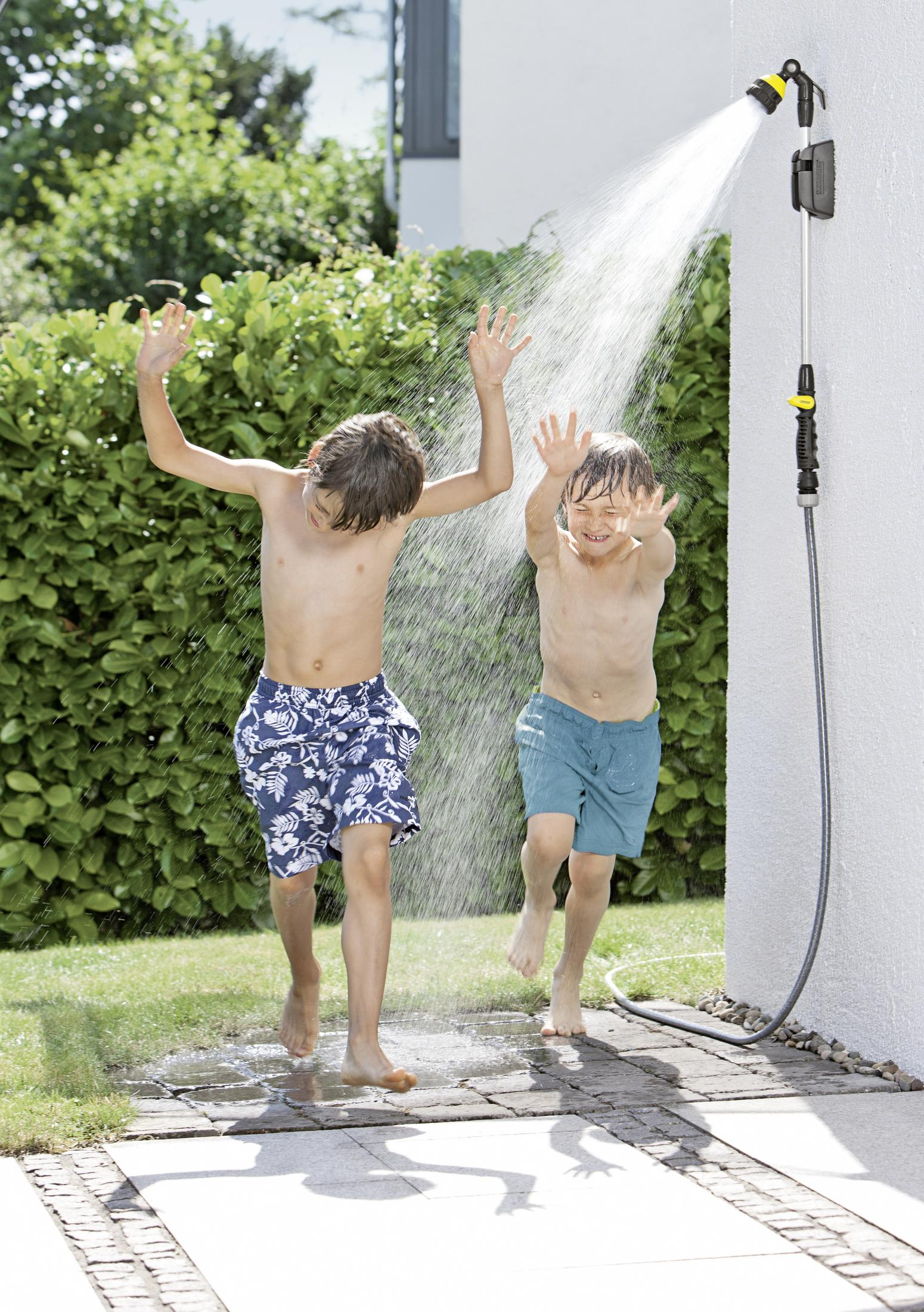 Zwei Jungen in Badehosen rennen durch Wasser aus einem Duschkopf.