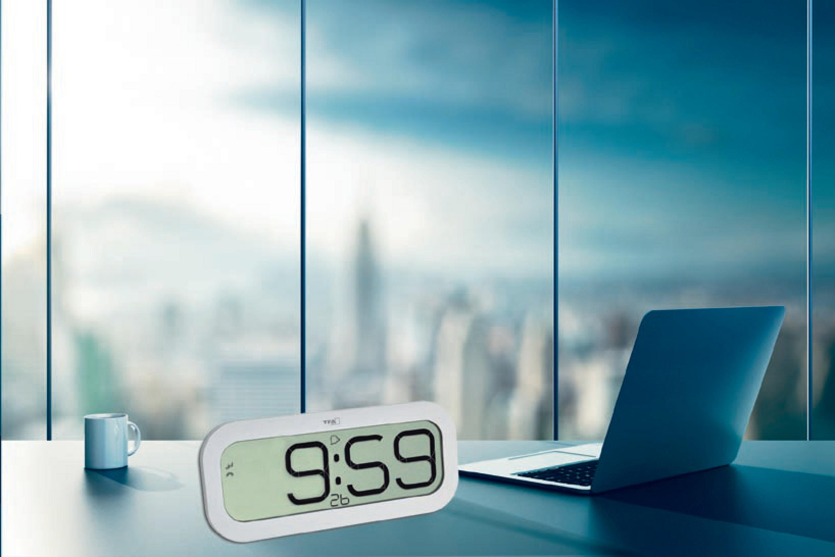 Orologio digitale che mostra le 9:59, accanto a un laptop e una tazza. Finestre sullo sfondo.