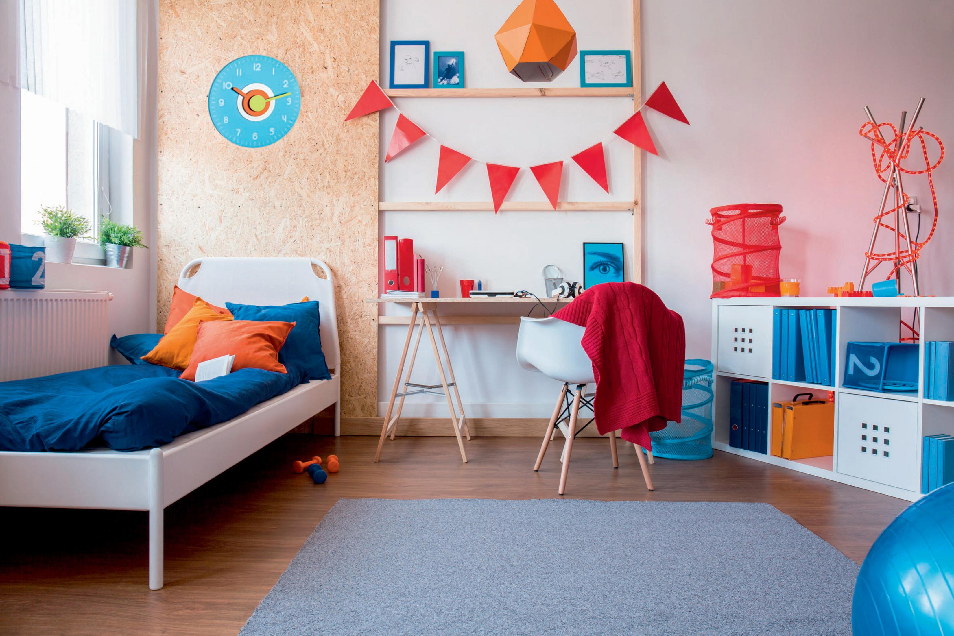 Ein farbenfrohes Kinderzimmer. Es gibt ein Bett, einen Schreibtisch und Regale. Dekorationen umfassen eine Wanduhr und Flaggen.