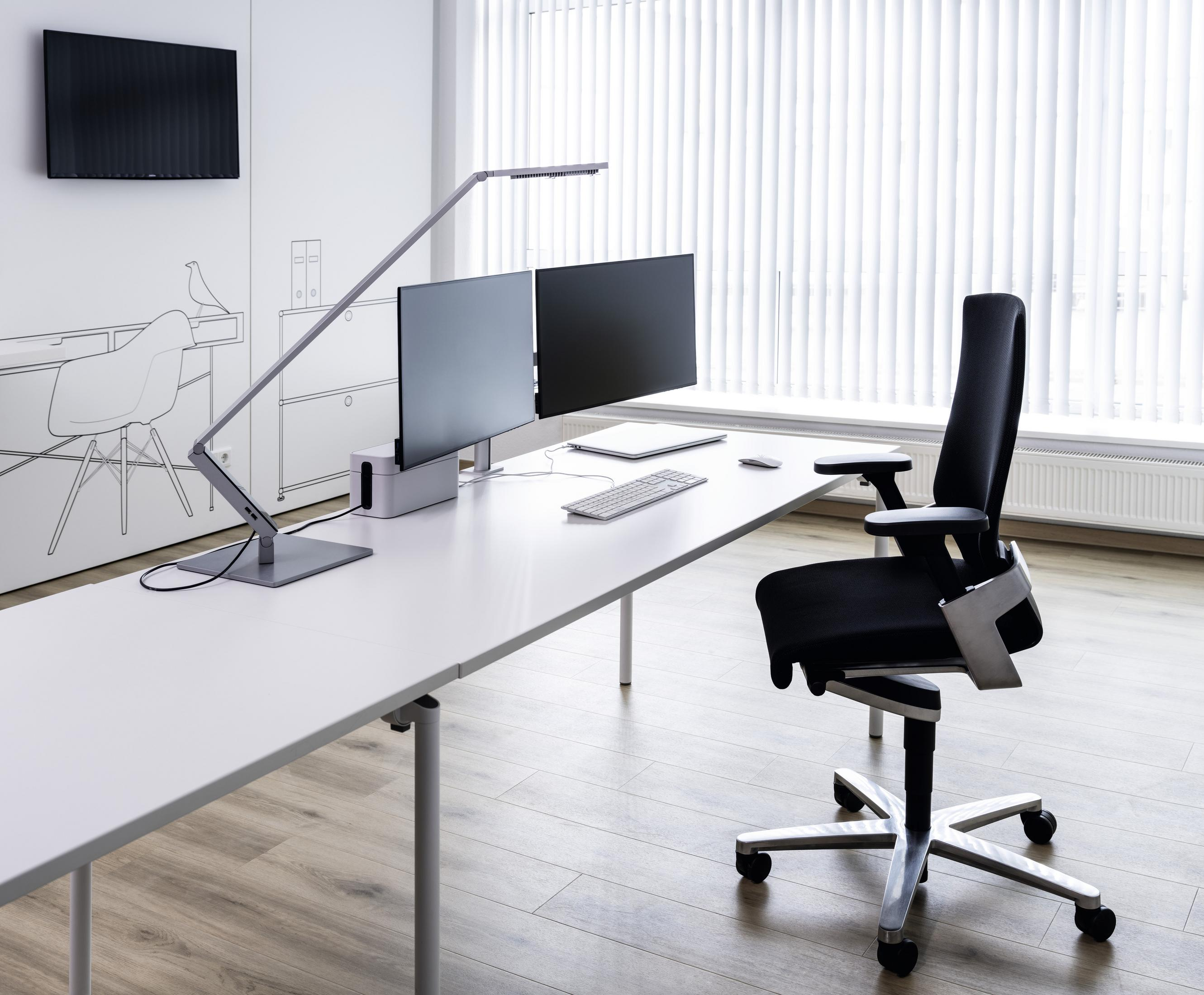 Ein Büroumgebung mit einem Schreibtisch, zwei Computermonitoren und einem ergonomischen Stuhl. Der Hintergrund ist hell.