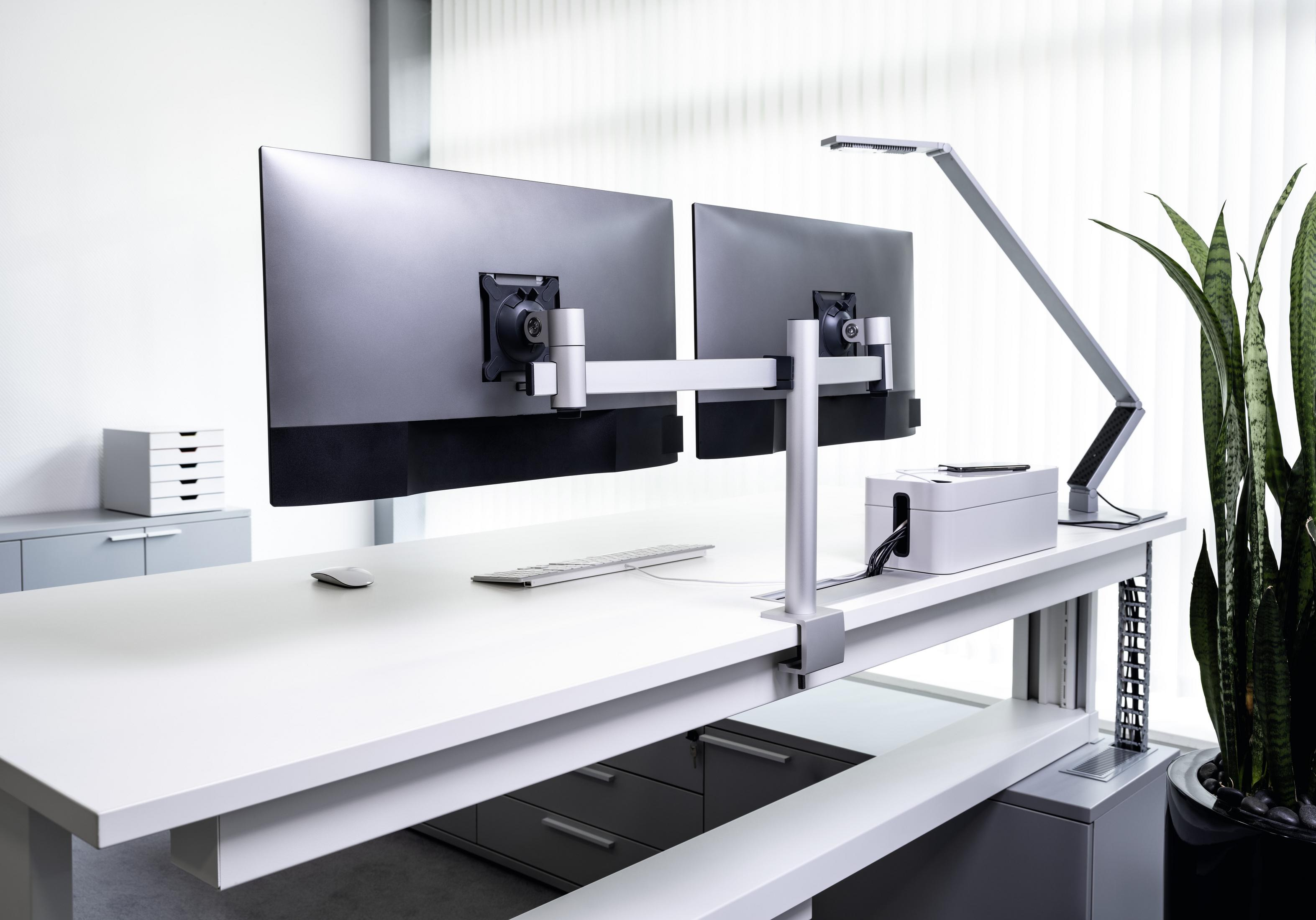 Ein weißer Schreibtisch mit zwei Monitoren. Eine Schreibtischlampe und ein Kabelorganizer sind auf dem Tisch.