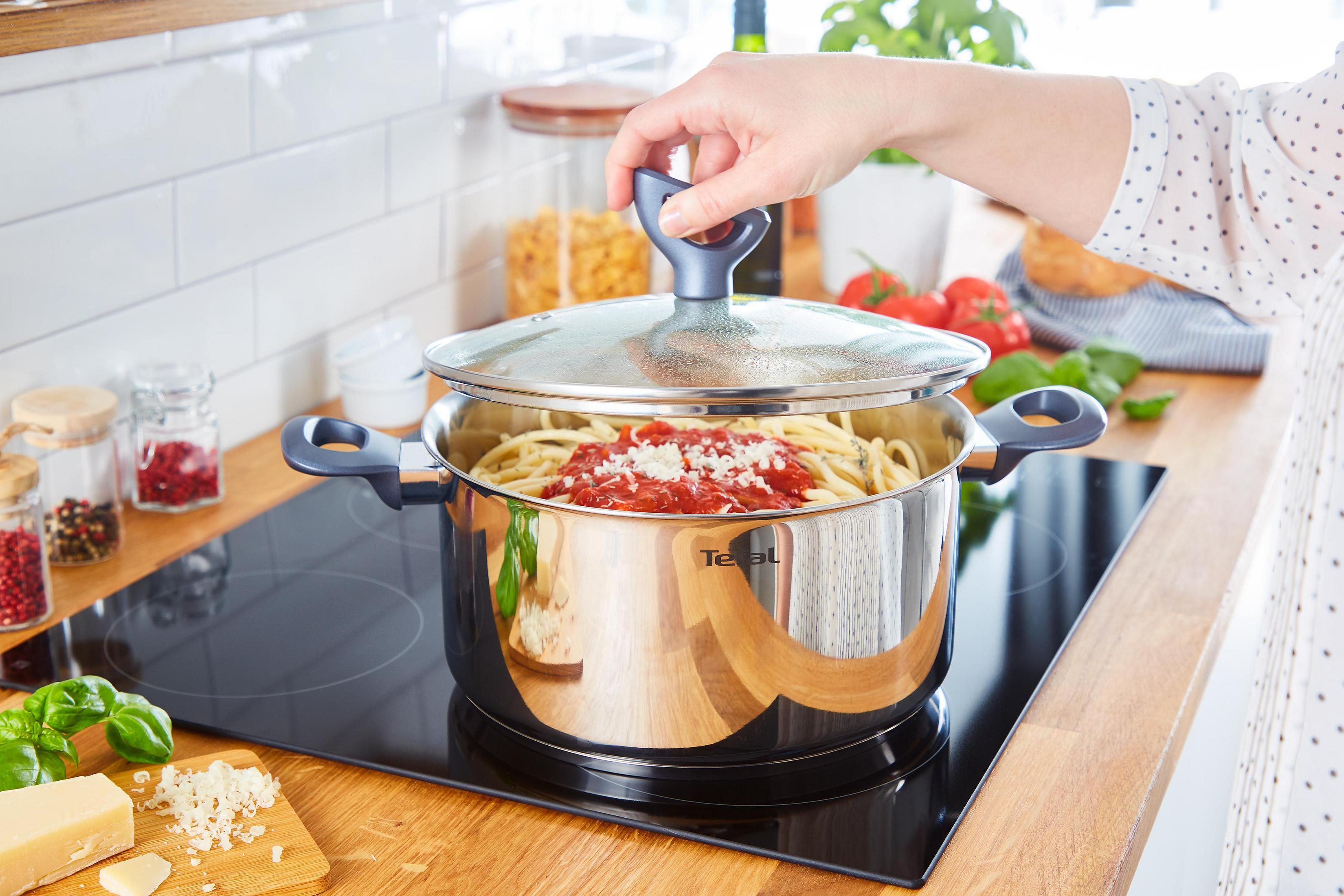 Frau hebt Deckel von Topf mit Spaghetti und Tomatensauce, auf Herdplatte. Küchenumgebung.