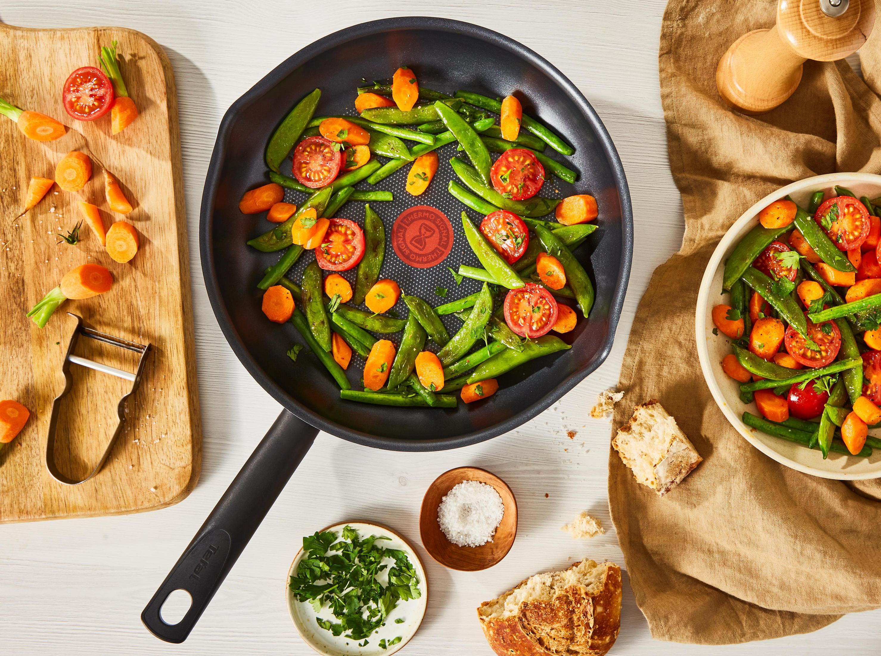 Een pan met gekookte groenten, waaronder erwten, wortelen en tomaten, met ingrediënten en een kom.