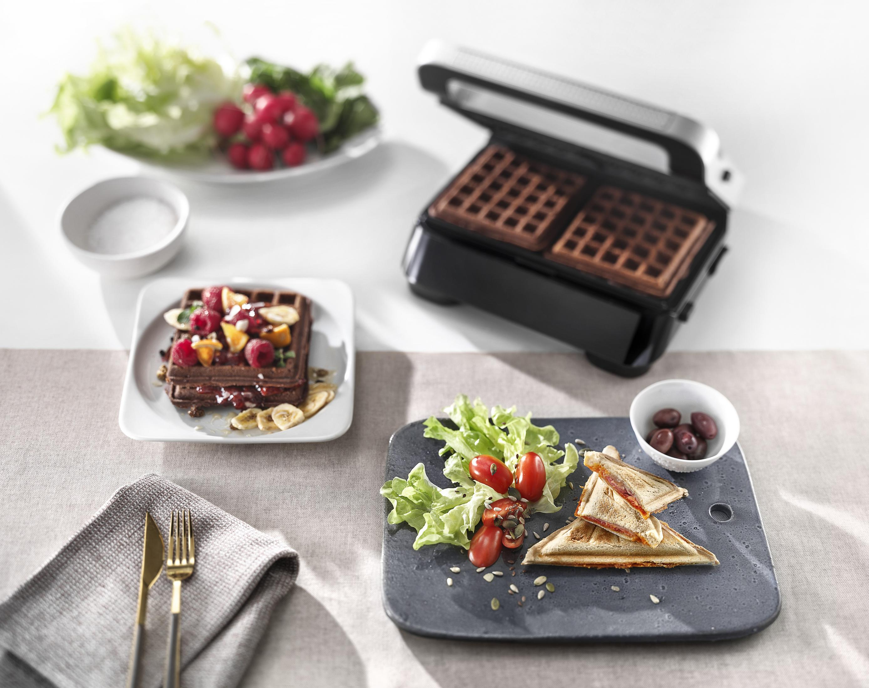 Una gofrera con gofres, un plato con gofres y coberturas, y otras comidas en una mesa.