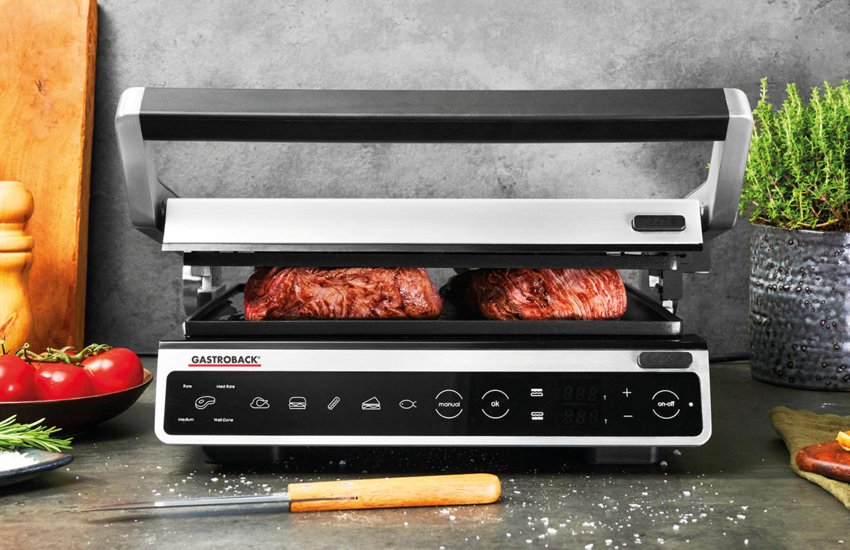 Un grill noir et argent avec deux steaks cuisant à l'intérieur, avec une planche à découper en bois et un couteau.