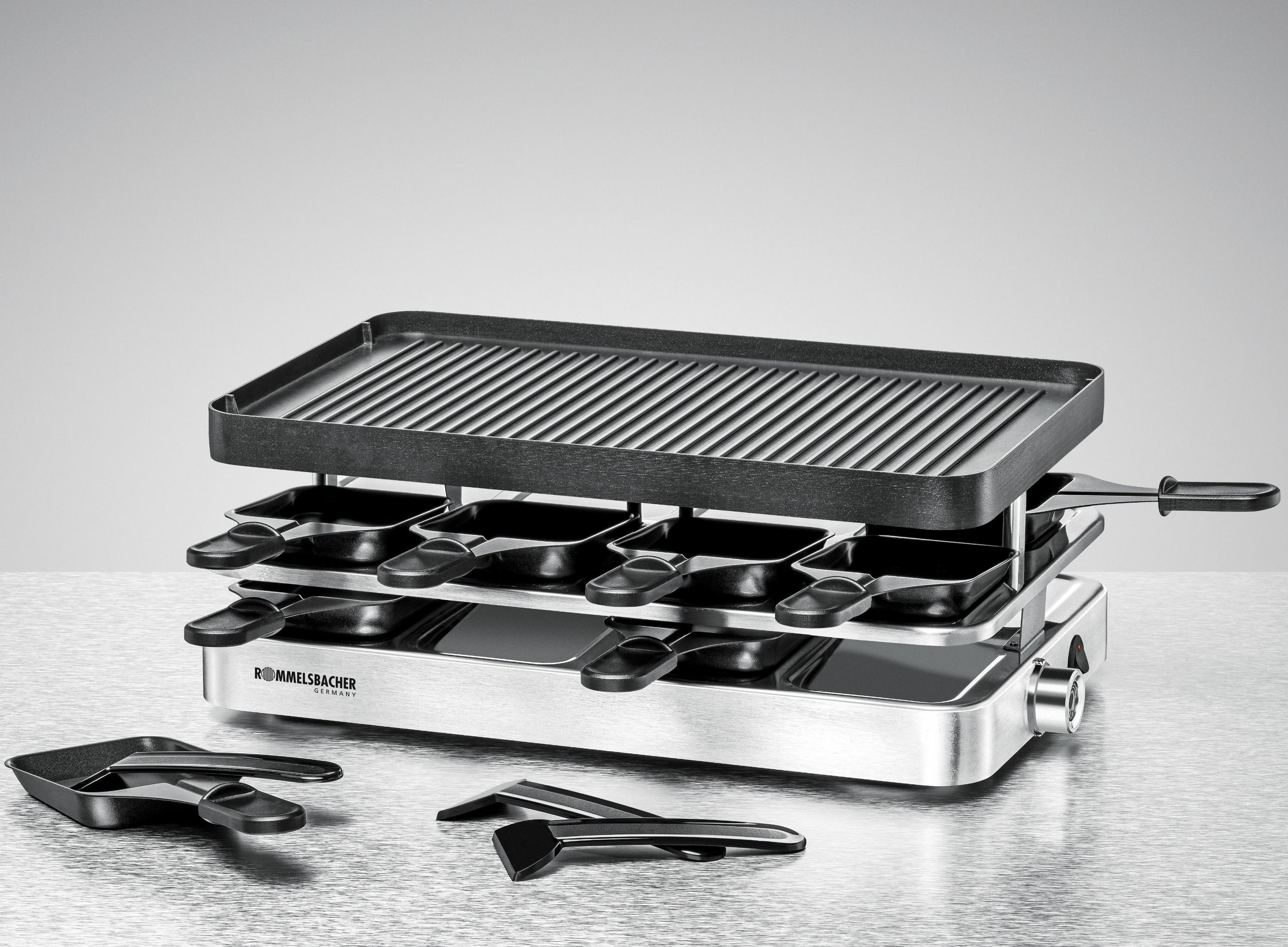 Grill do raclette z czarnym rusztem i patelniami poniżej. Srebrna podstawa. Białe tło.