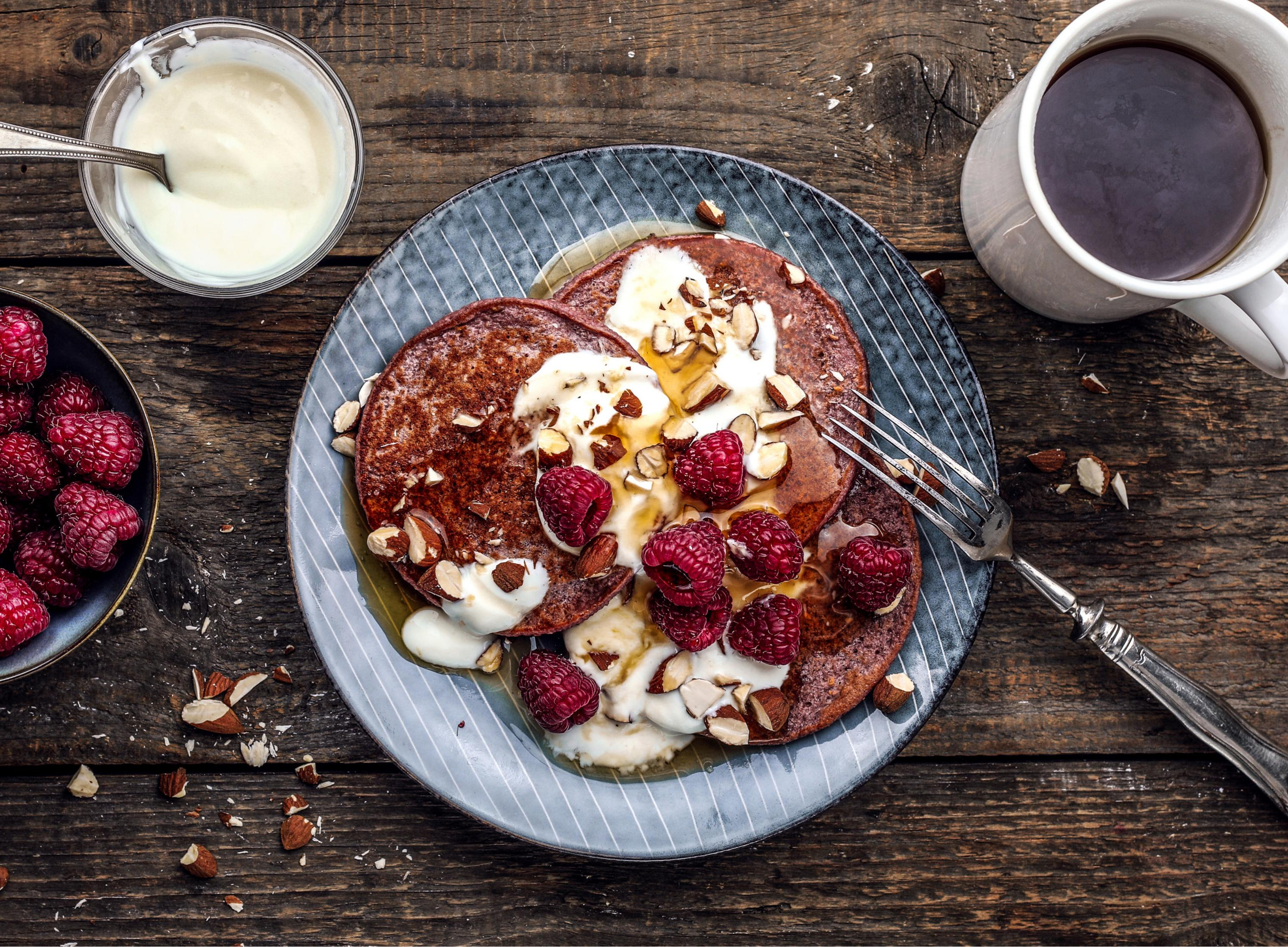 Naleśniki z dodatkami, malinami i filiżanką kawy na drewnianym stole. Miska jogurtu.