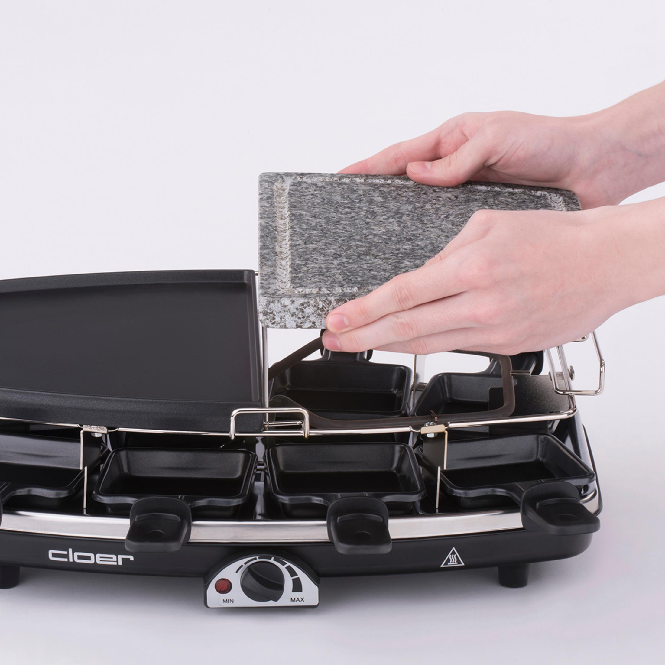 Hände, die eine Steinplatte auf einen Raclette-Grill legen. Schwarze Pfannen darunter. Weißer Hintergrund.