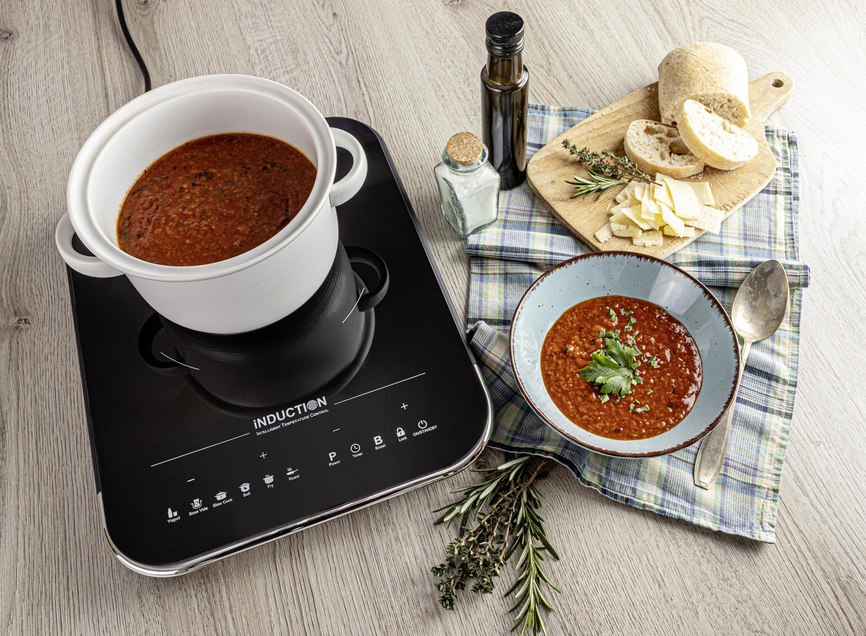 Biały garnek z sosem pomidorowym na płycie indukcyjnej. Miseczka z zupą, chlebem i ziołami.