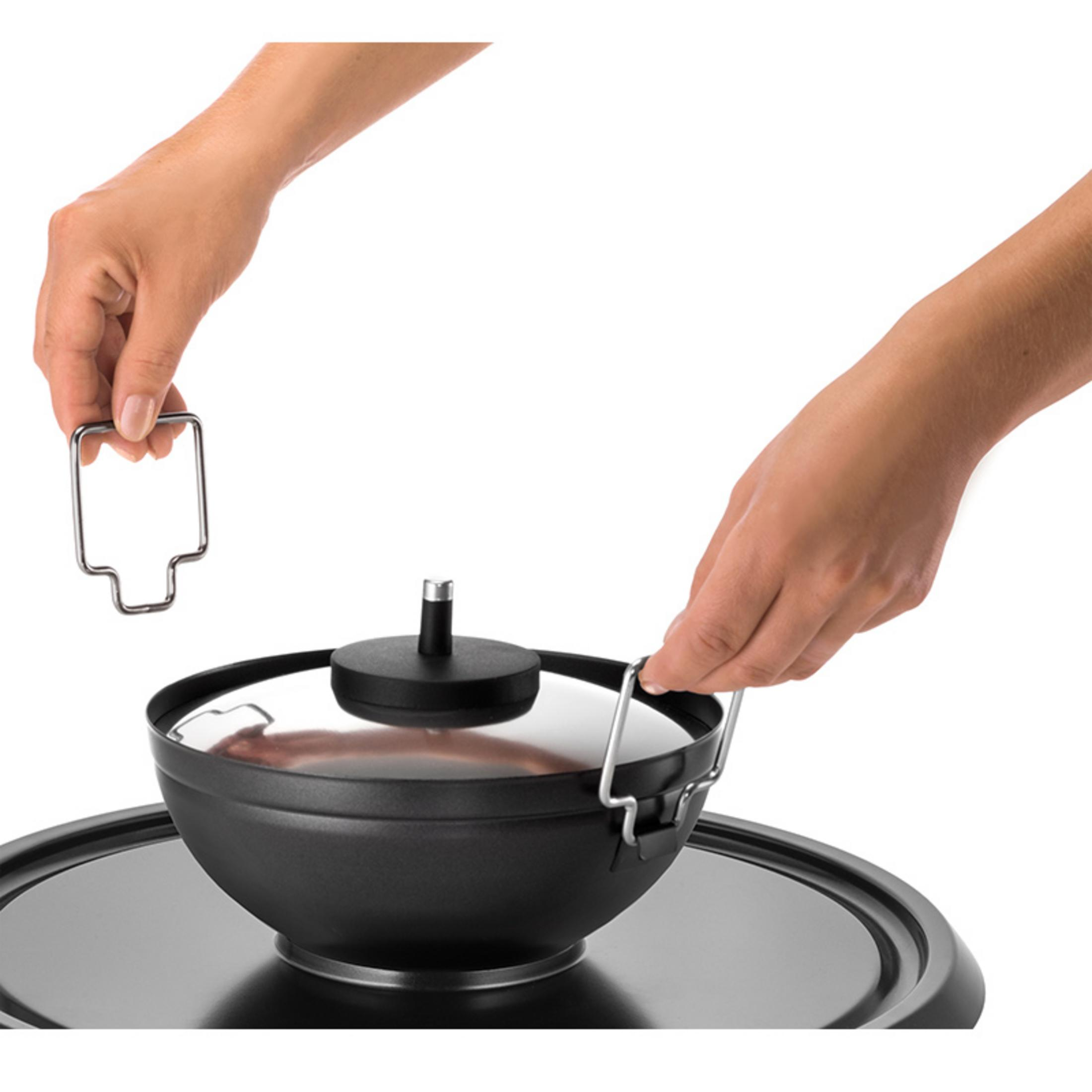Ręce trzymające uchwyty czarnego garnka z błyszczącą pokrywką, na czarnej powierzchni.