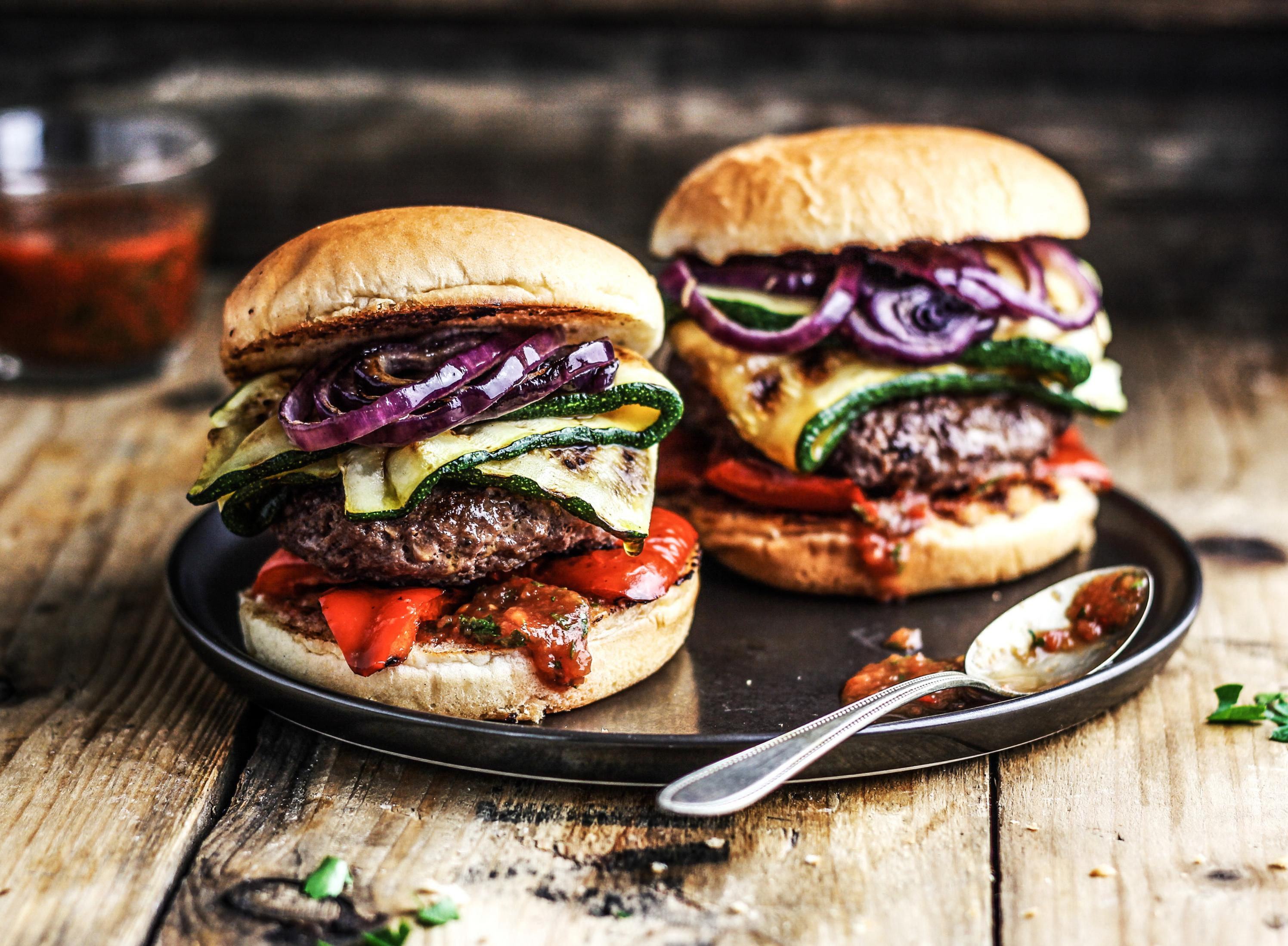 Dwa burgery na talerzu z dodatkami: cukinią, czerwoną cebulą i sosem pomidorowym.
