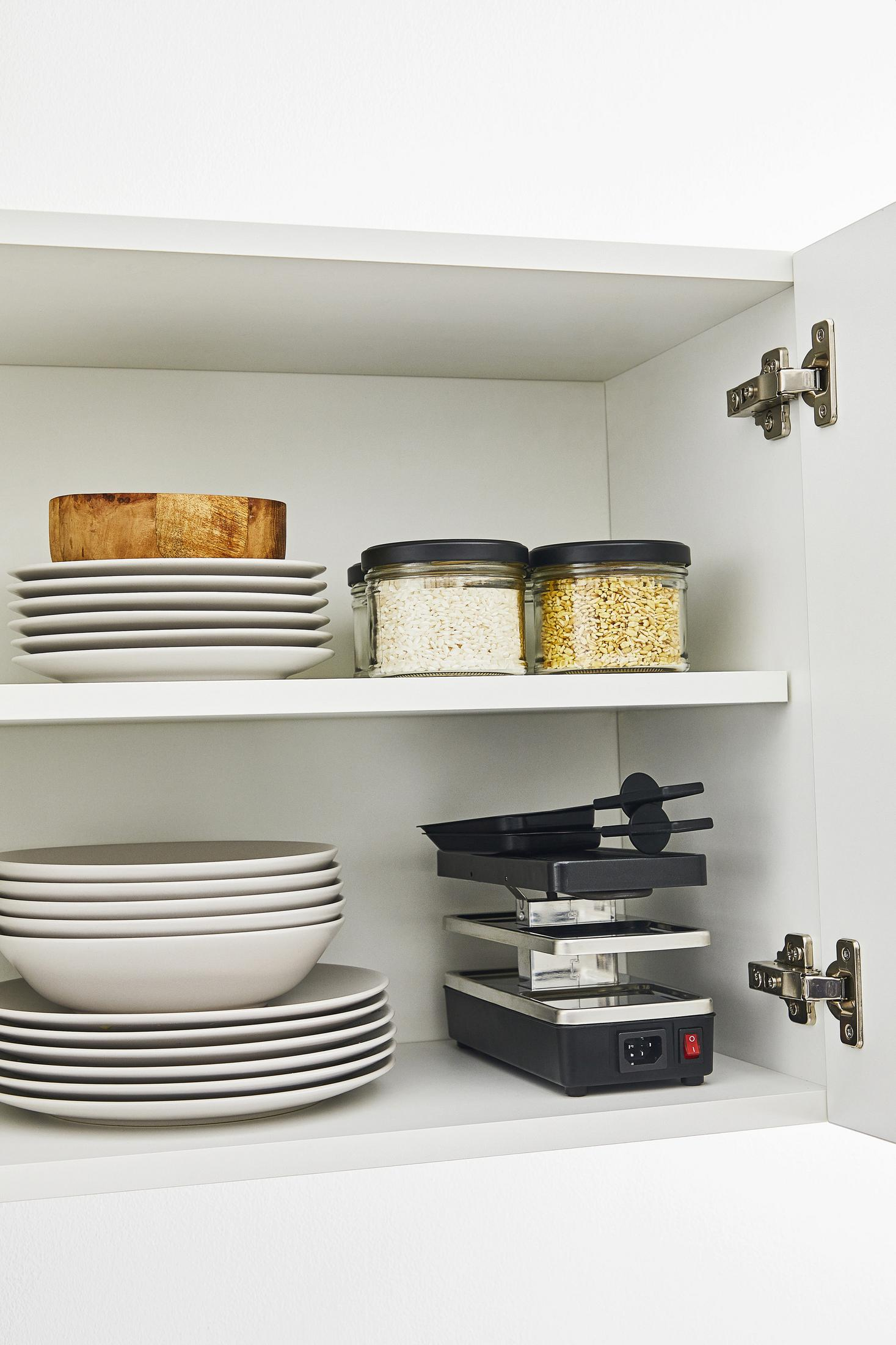 Biała szafka kuchenna z talerzami, miskami i słoikami z jedzeniem. Na dolnej półce grill do raclette.