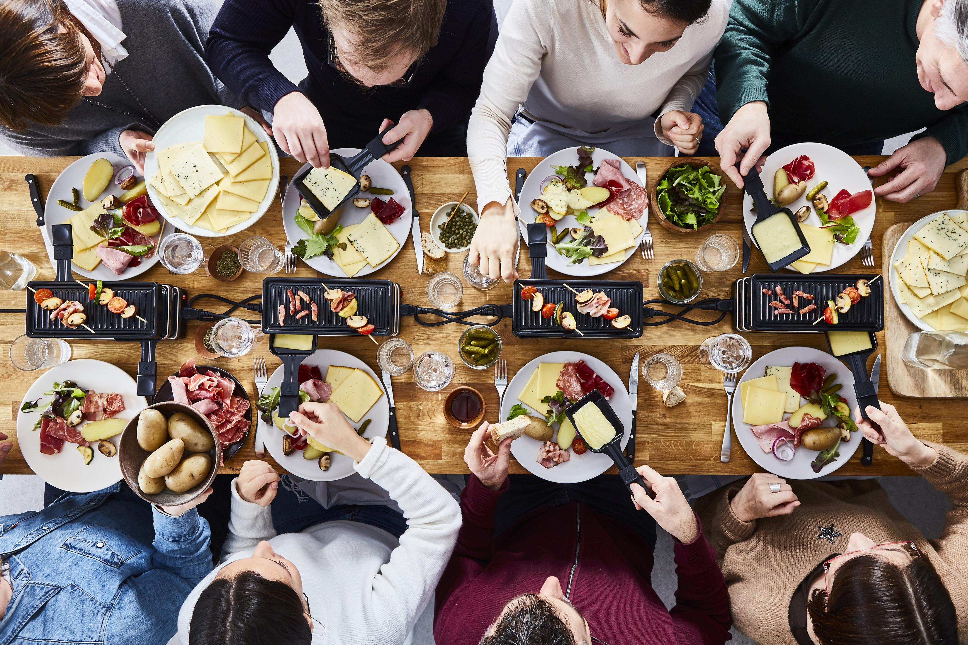 Grupa osób wokół stołu z grillami do raclette i jedzeniem. Ludzie jedzą i wchodzą w interakcje.