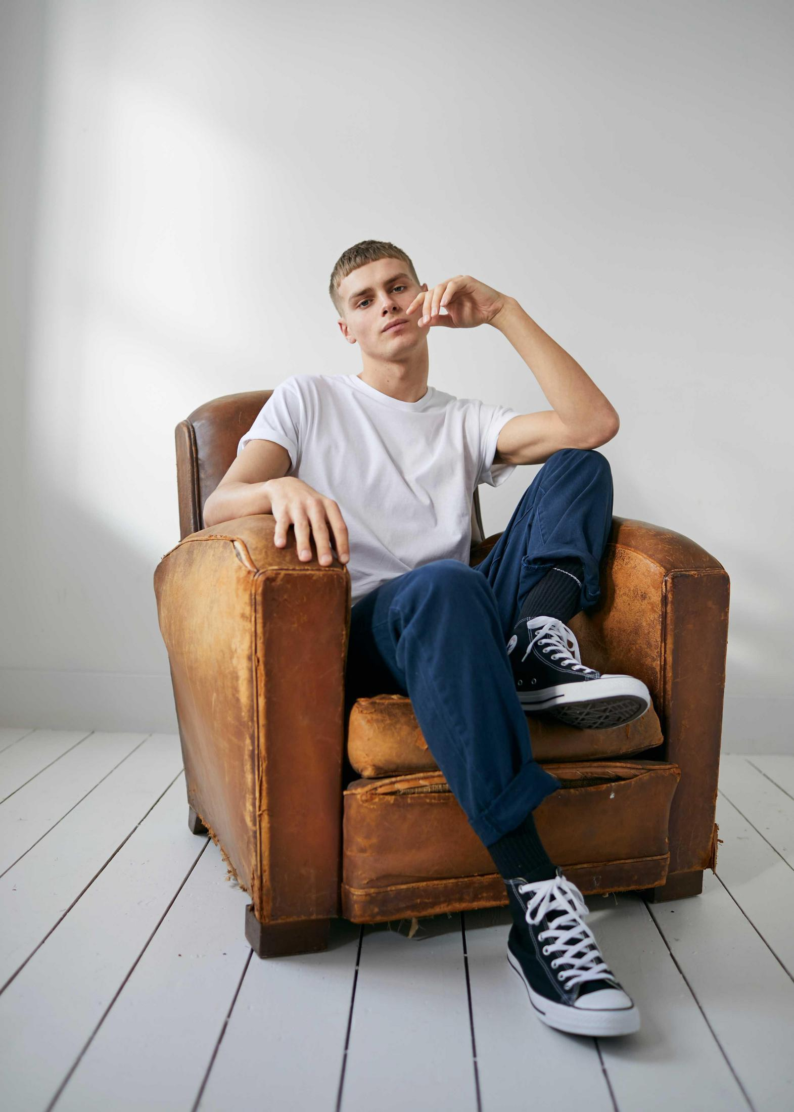 Młody mężczyzna siedzi w zużytym skórzanym fotelu, ubrany w biały t-shirt, dżinsy i czarne trampki.
